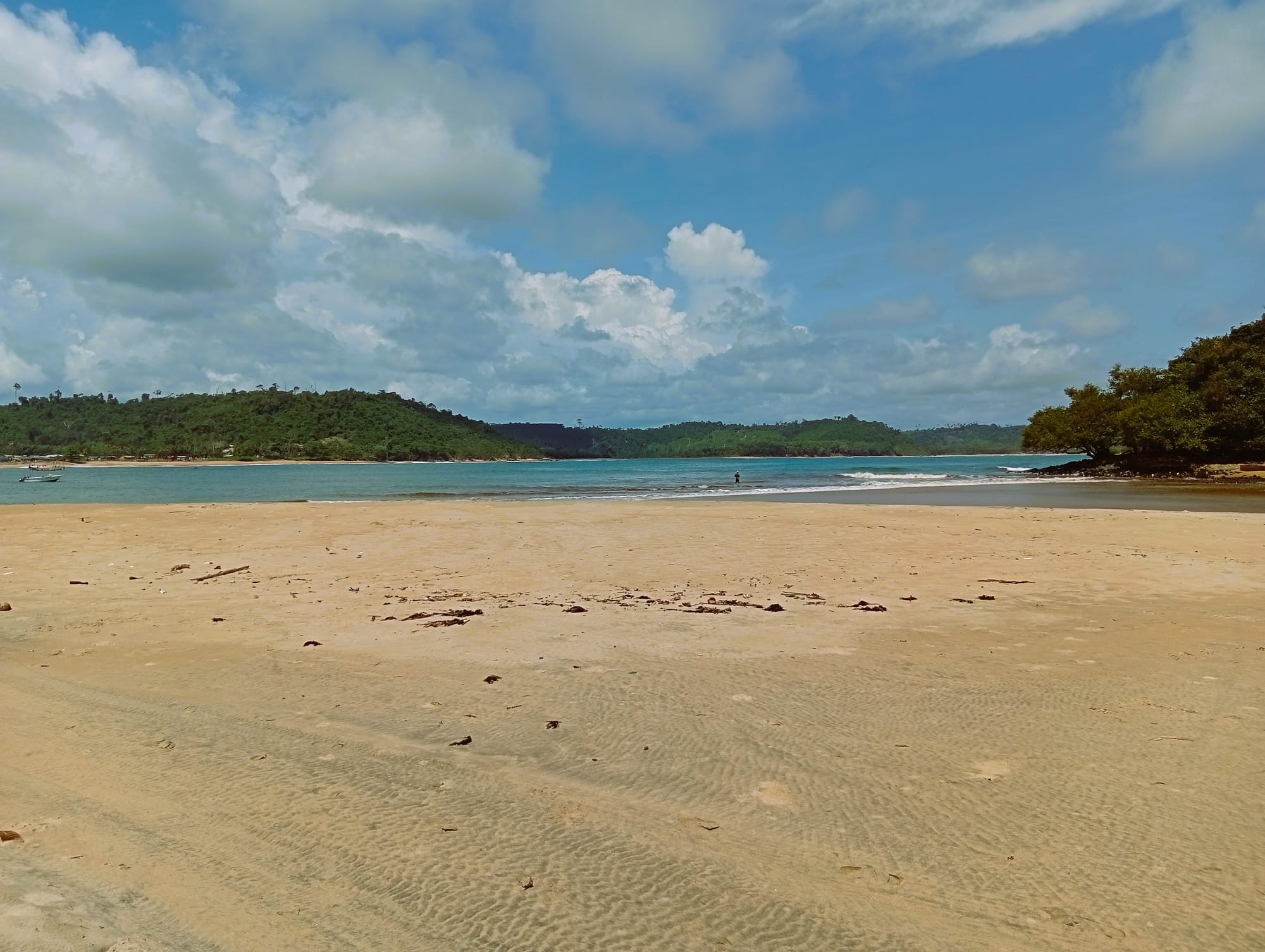 A wide, empty sandy beach curving round a turquoise bay backed by low forested hills under a blue sky with white clouds