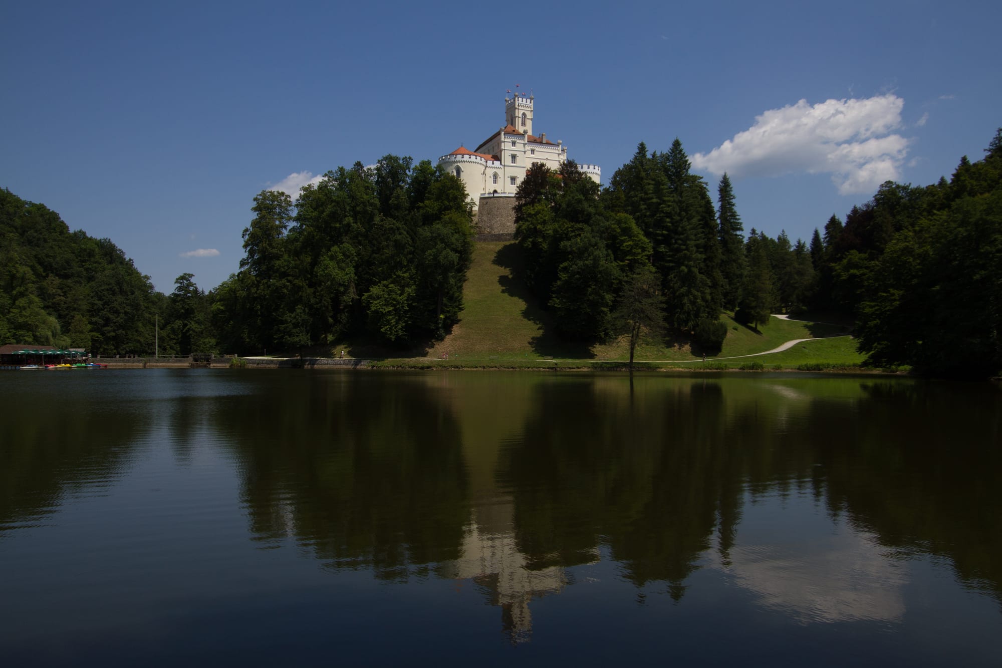 White hilltop Trakošćan Castle above wooded slopes, reflected in the still water of its lake under a blue sky