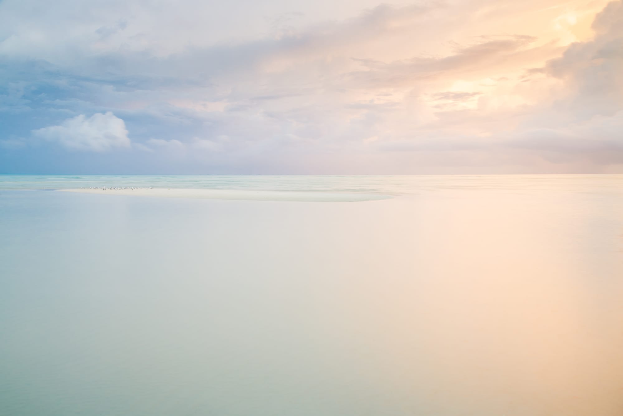 Calm, shallow turquoise water and a pale sandbar under soft pink and grey clouds at sunrise