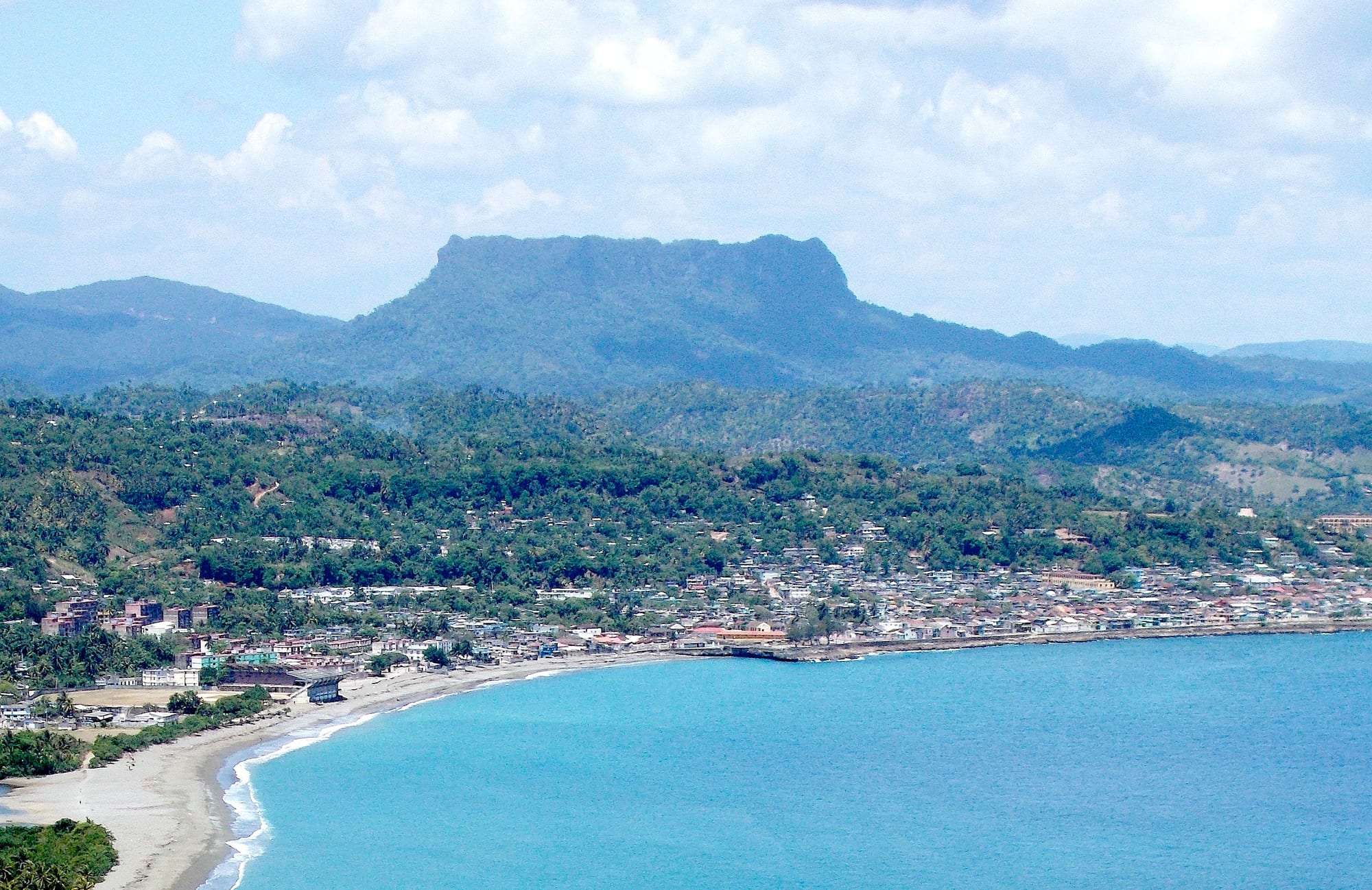 A curving bay of blue water and a sandy beach below the town of Baracoa, with a flat-topped forested mountain behind