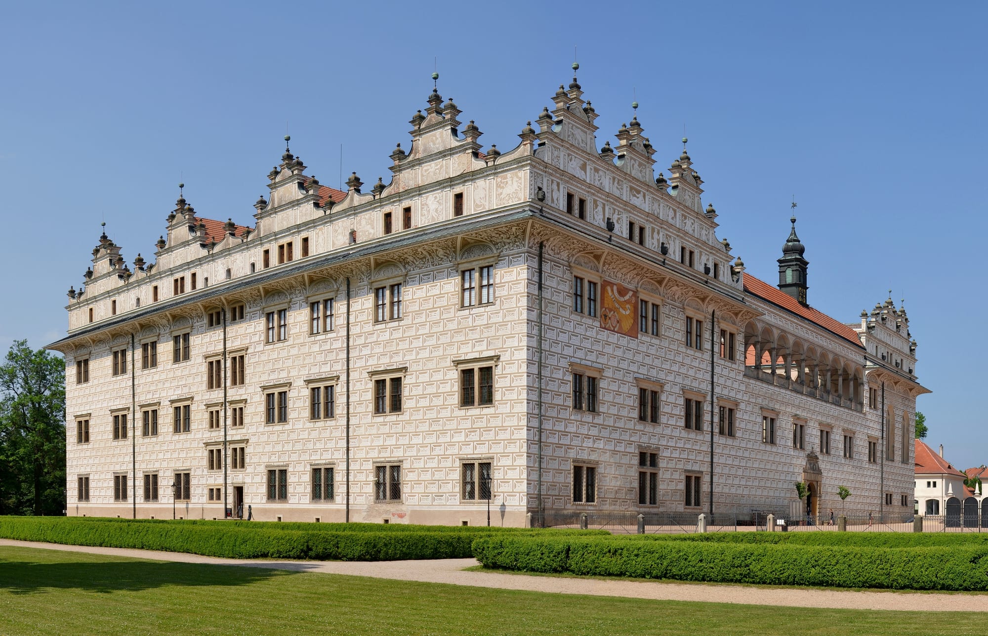 A long Renaissance chateau covered in pale sgraffito patterning, with ornate gables and an arcaded wing, behind clipped hedges and a lawn
