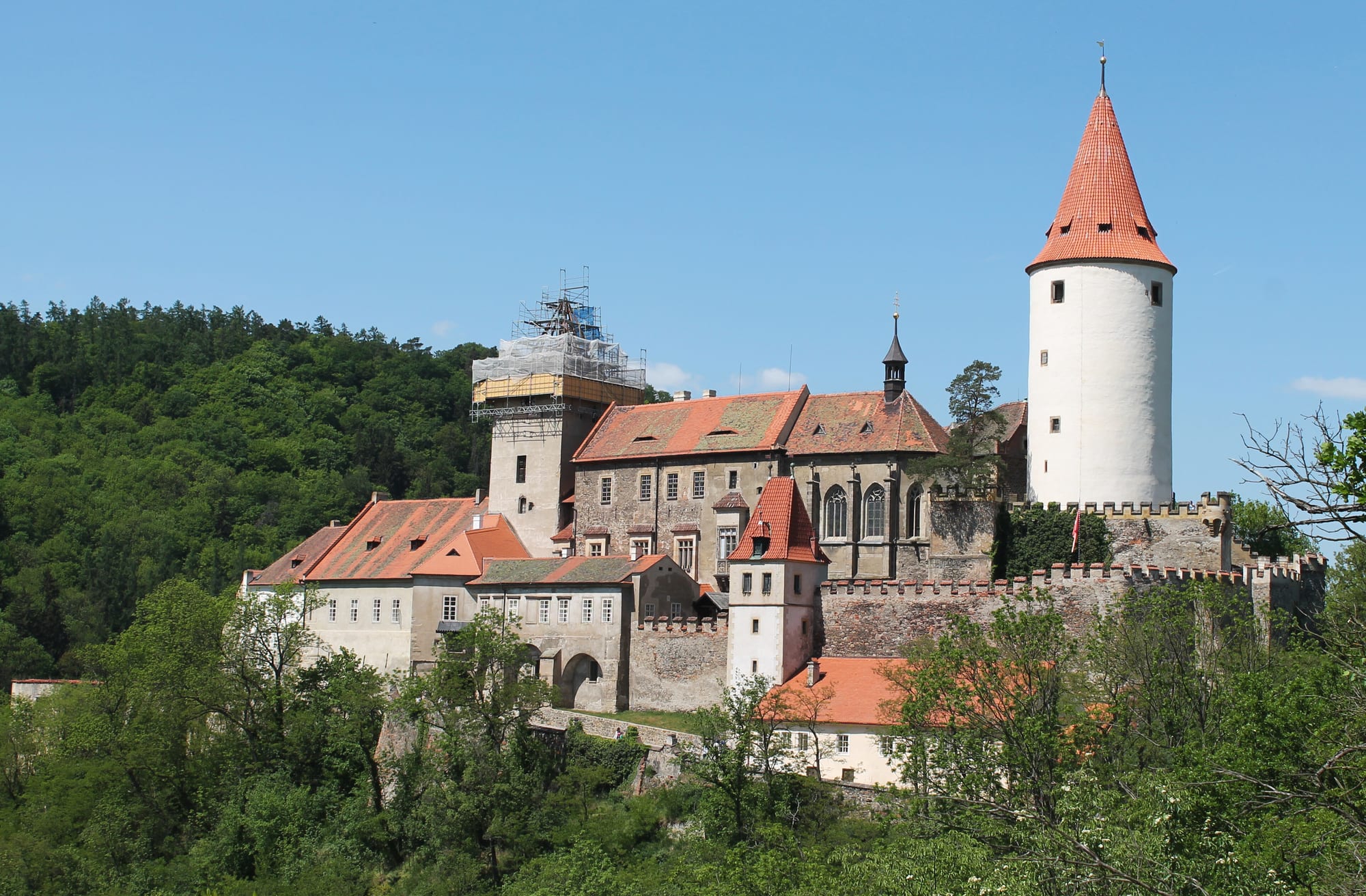 A medieval castle with red-tiled roofs, battlemented walls and a tall white round tower, set among thickly wooded hills, with one tower under scaffolding