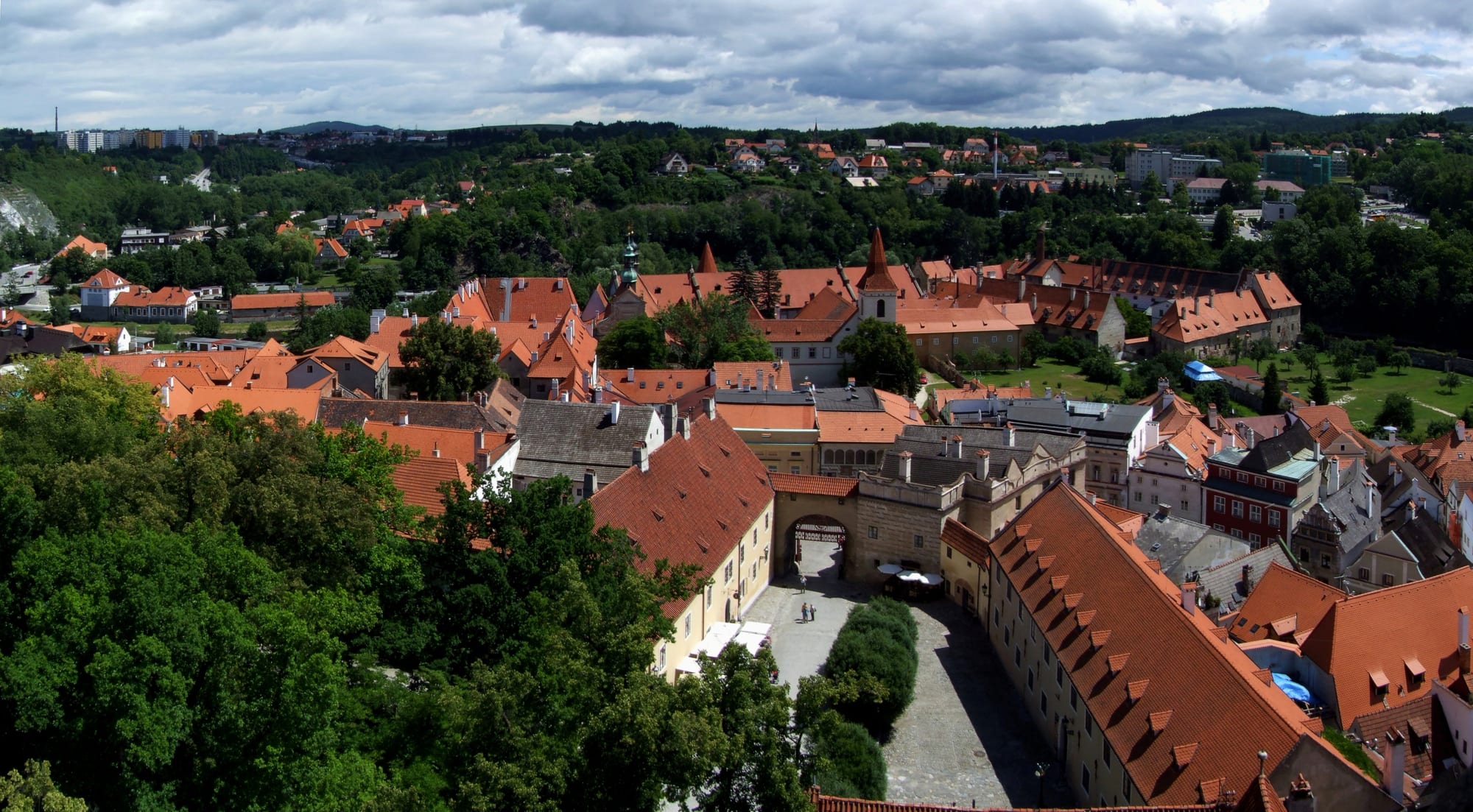 Red-tiled roofs, a church spire and a cobbled courtyard with an arched gateway, surrounded by trees and green hills