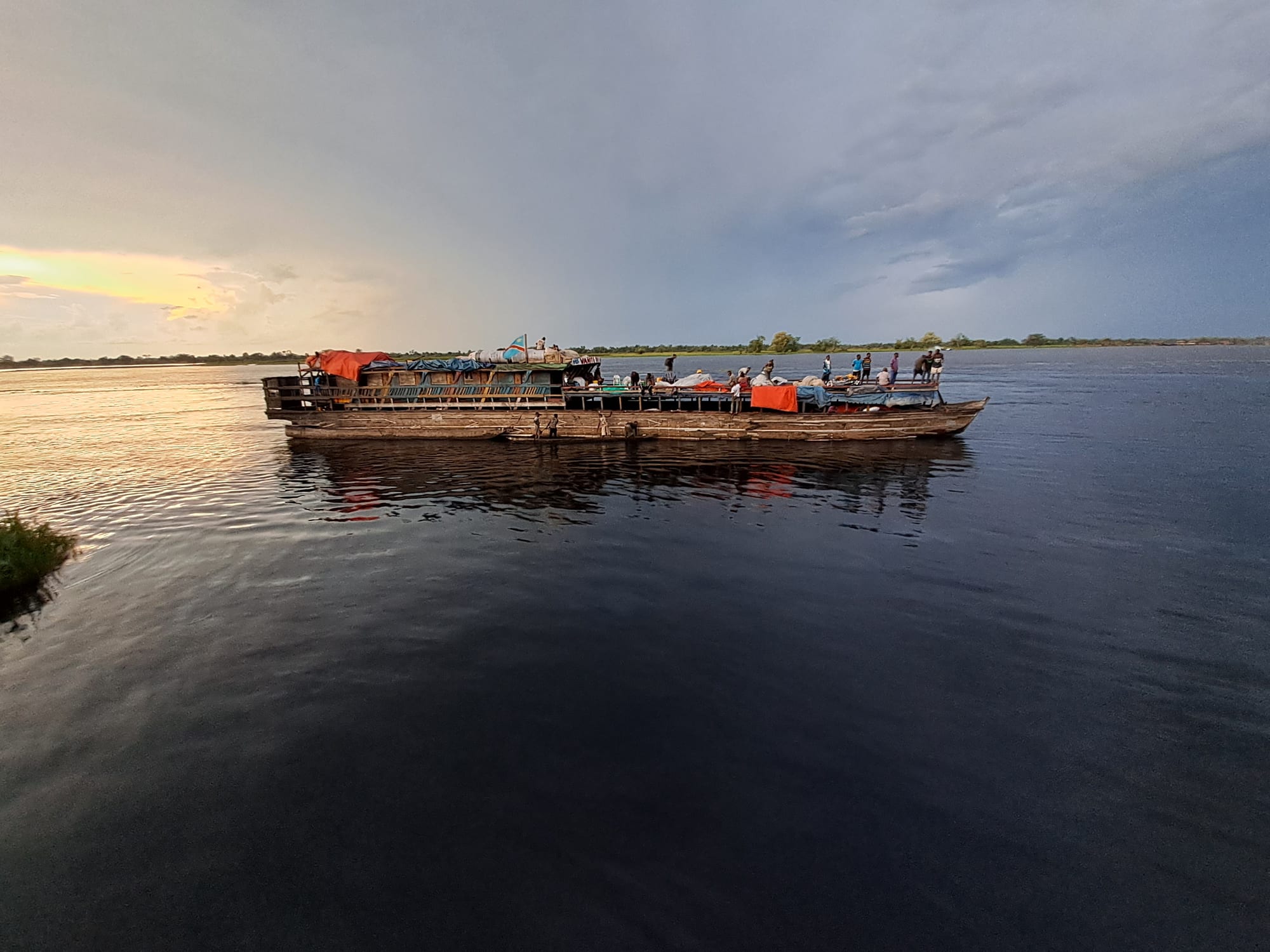 A loaded wooden river boat with passengers on deck crossing a wide, calm river under a grey evening sky