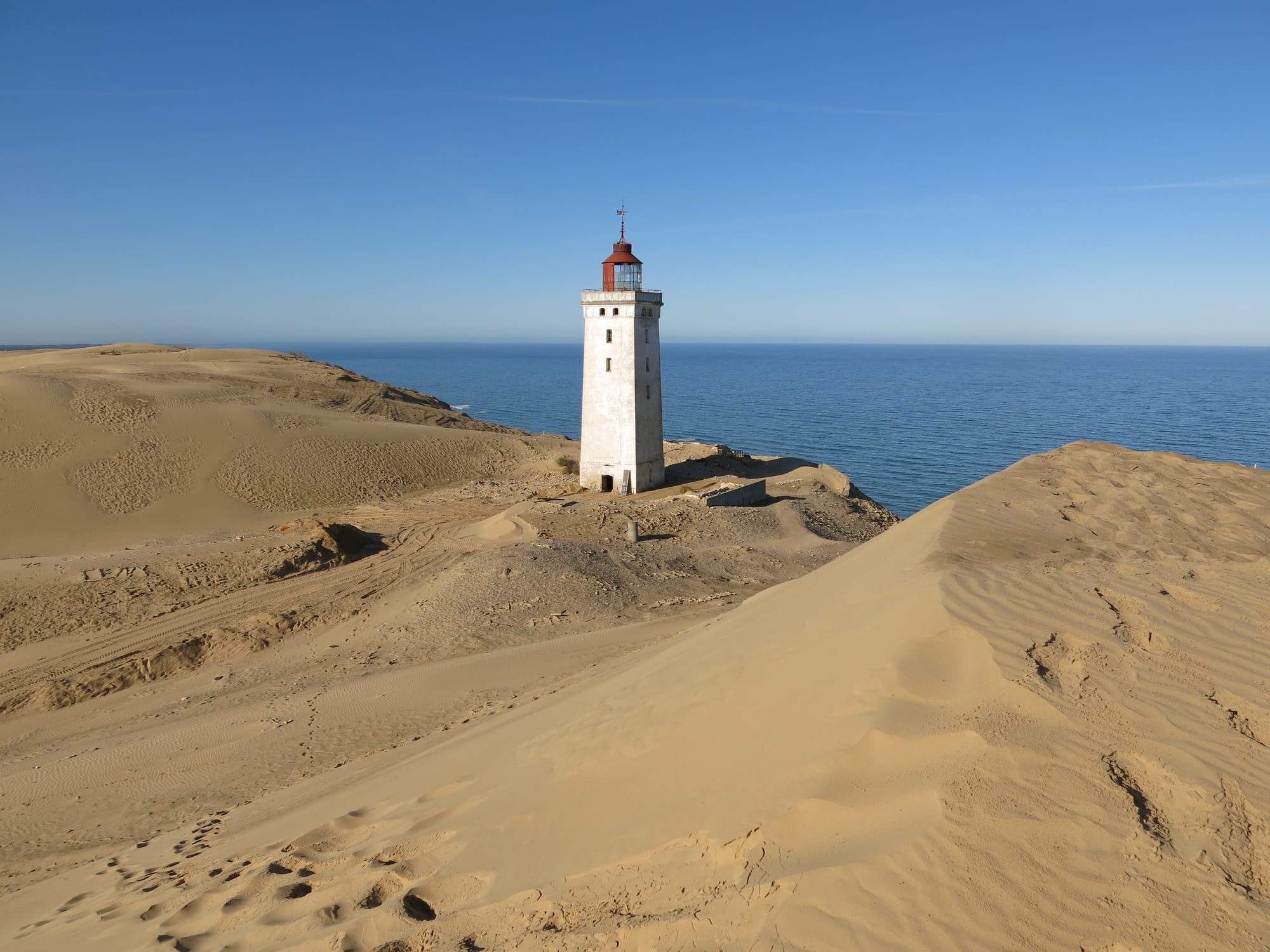 A white lighthouse with a red lantern standing alone among bare golden sand dunes above a calm blue sea