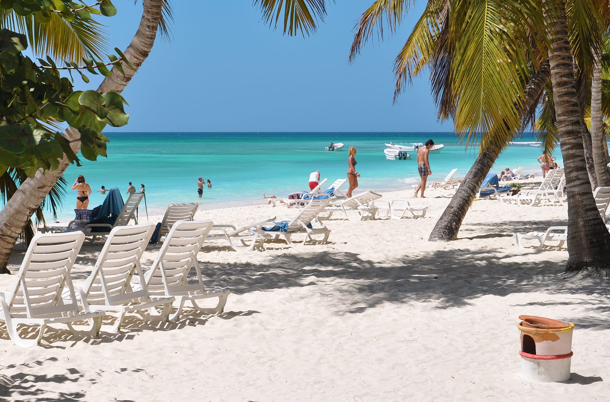 White sand beach with sun loungers under palm trees, facing clear turquoise water with boats moored offshore