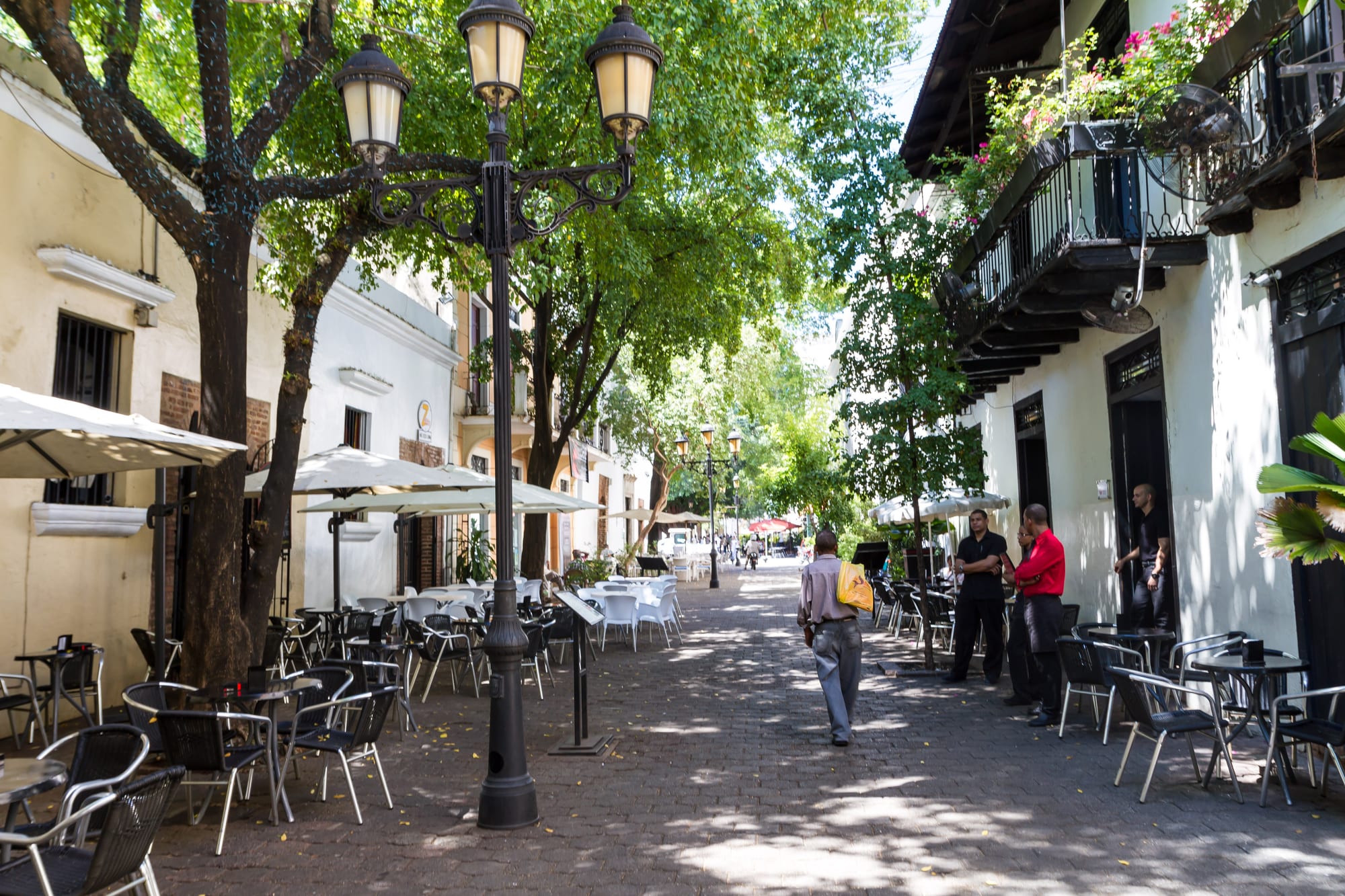 A shaded pedestrian street of pale colonial houses with café tables, iron lamp posts and balconies with flowers