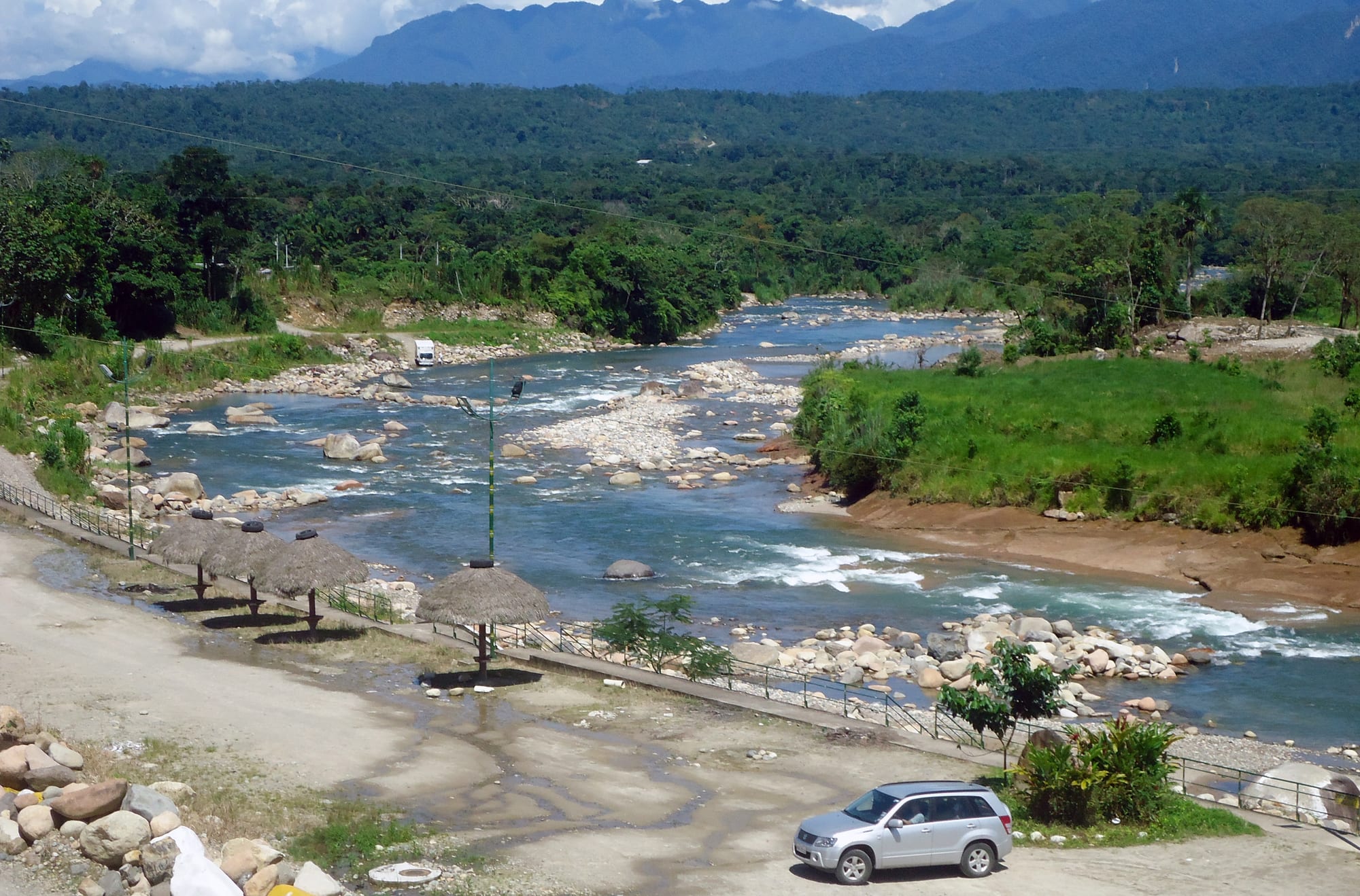 A clear, rocky river winding through green lowland forest, with thatched shelters on the bank and forested mountains behind