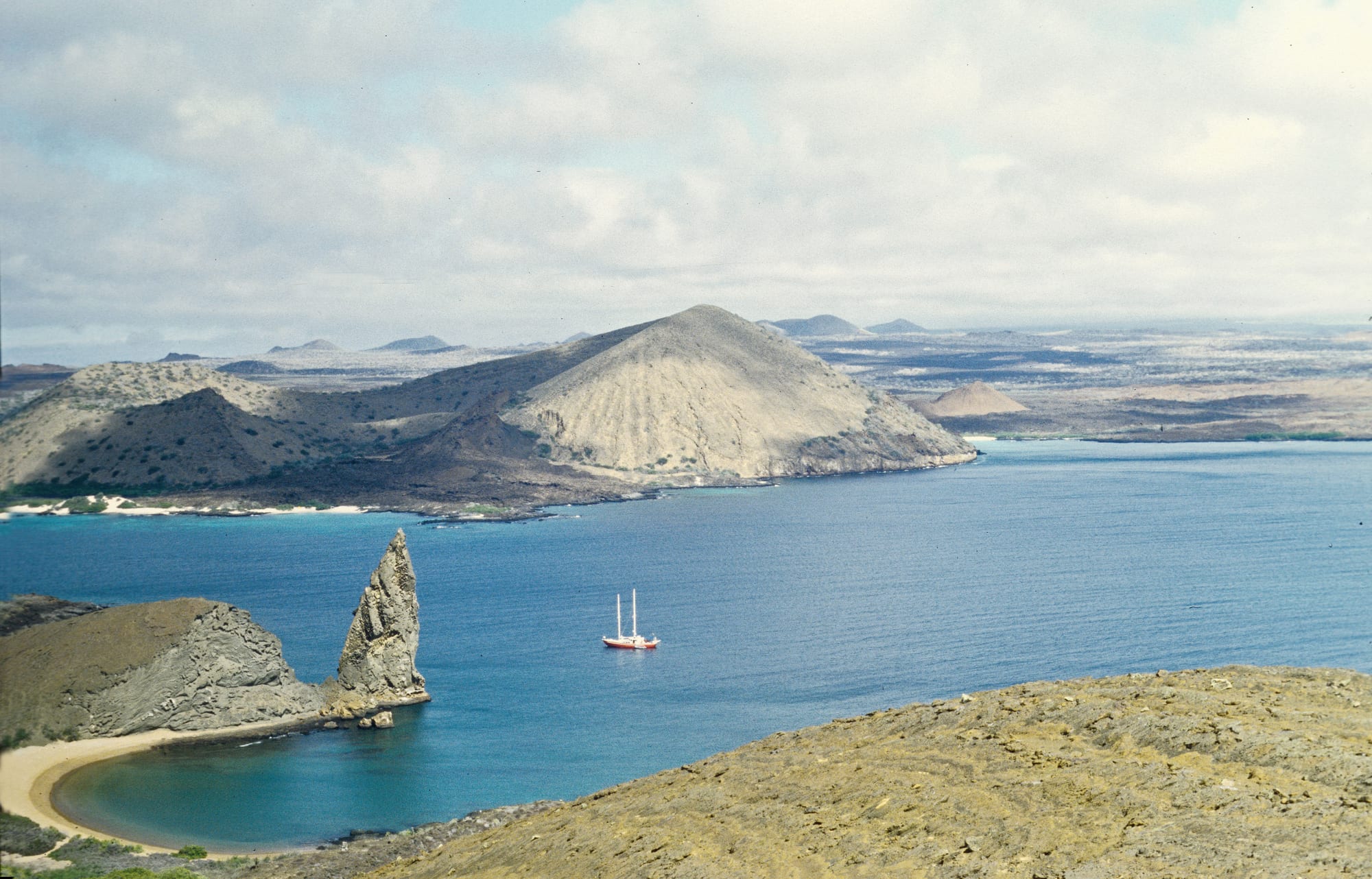 A pointed rock spire beside a curved sandy beach and blue bay, with bare volcanic hills and a sailing boat beyond