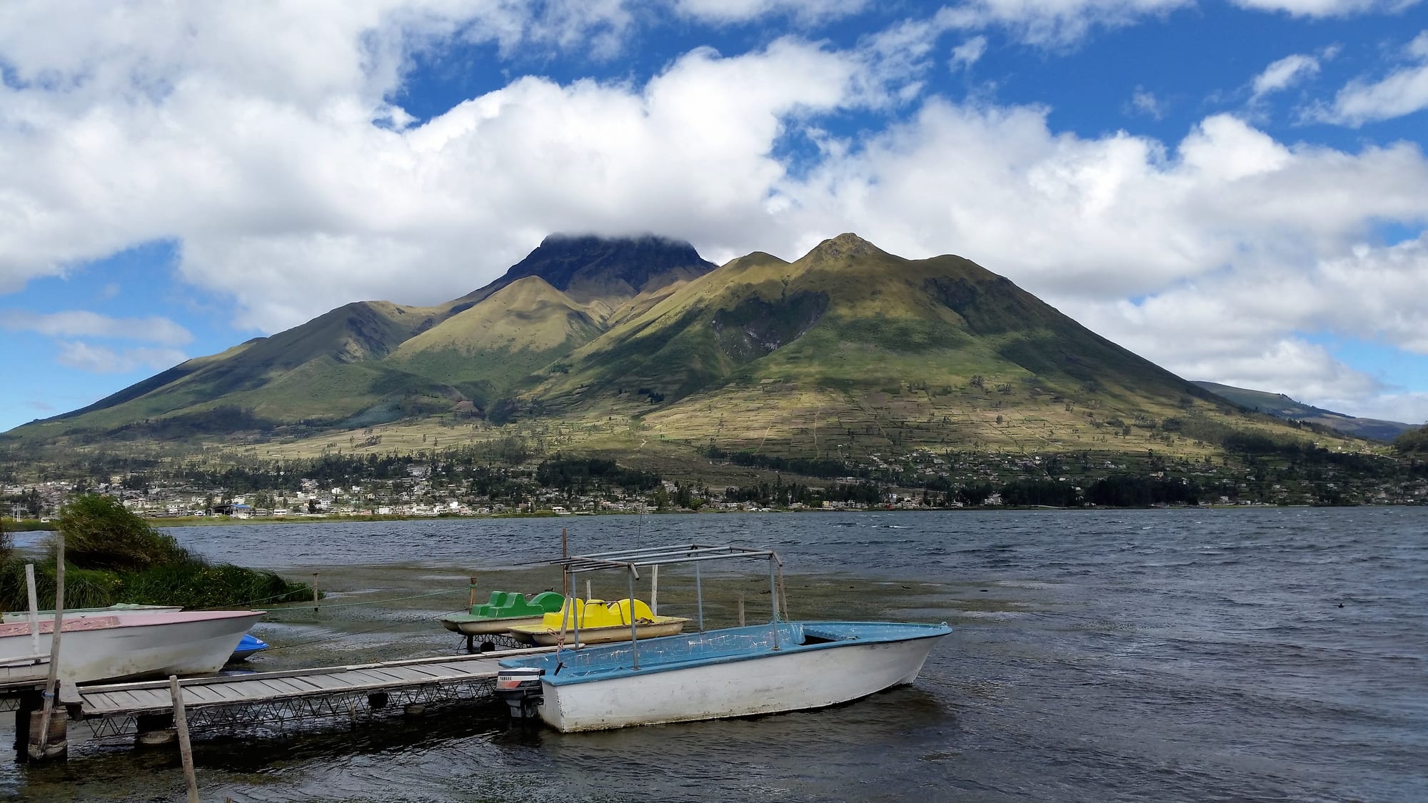 Small boats moored at a wooden jetty on a lake, with a green volcano and a lakeside town behind under white clouds