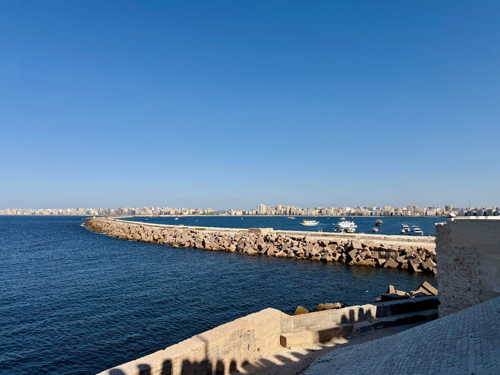 A stone breakwater running across deep blue water, with Alexandria's long line of seafront buildings behind