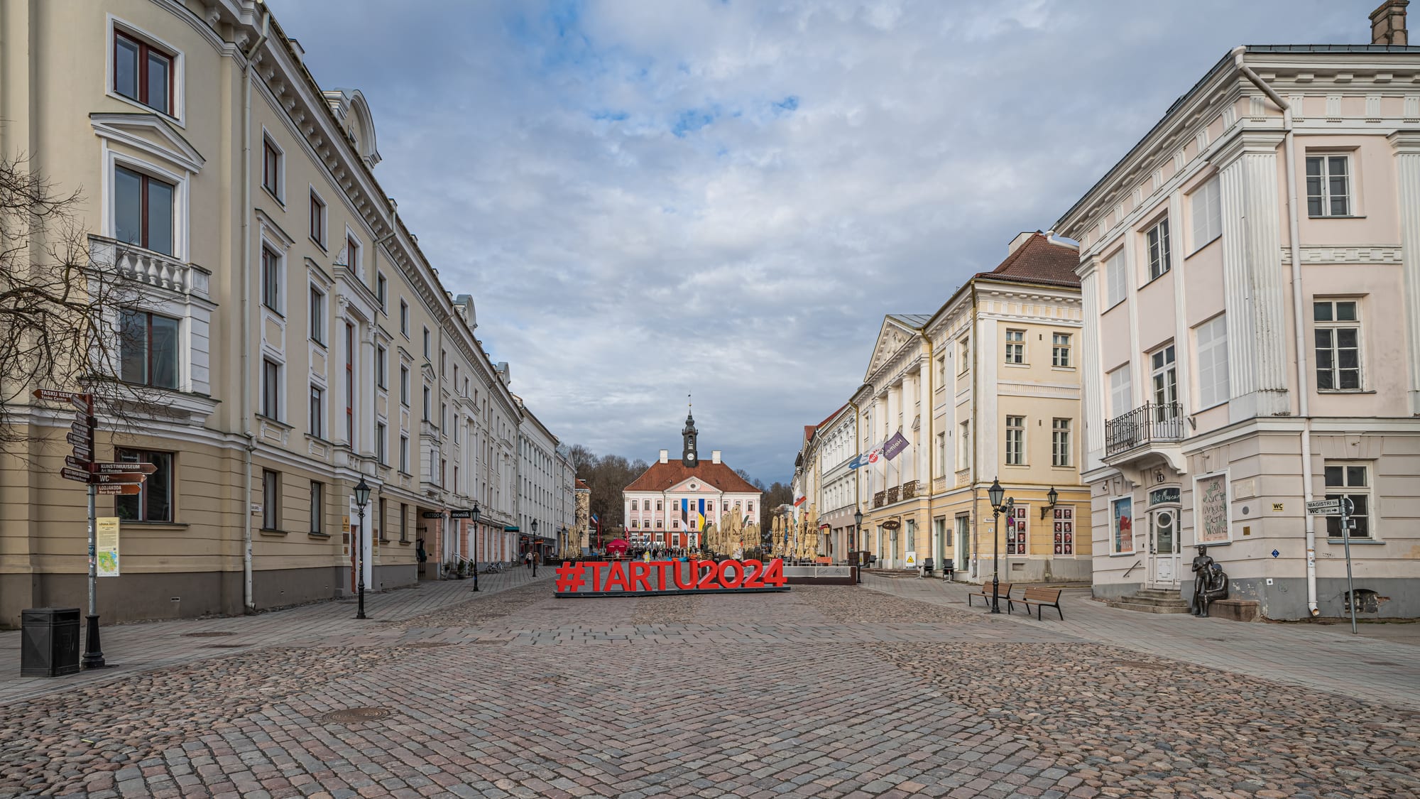 Cobbled Town Hall Square in Tartu lined with pale classical buildings, the pink town hall and its spire at the far end