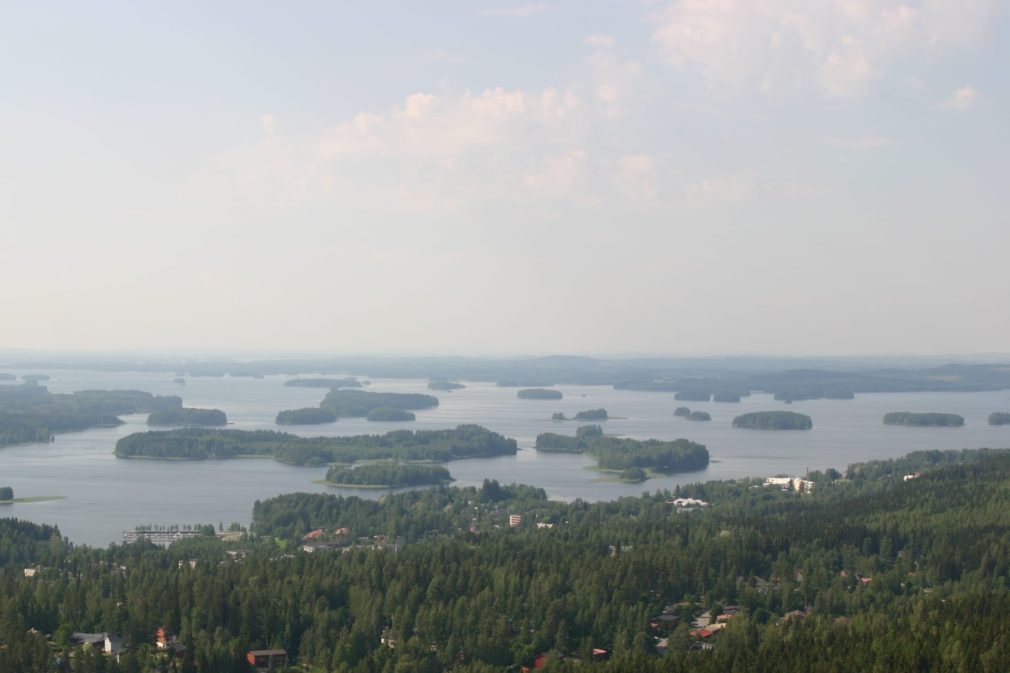 Forested islands scattered across a wide lake under a hazy summer sky, seen from a hill above pine forest and houses