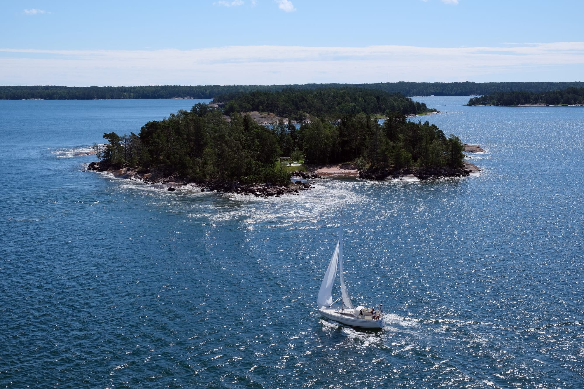 A sailing boat passing a small forested rocky island in sparkling blue sea, with more low wooded islands behind