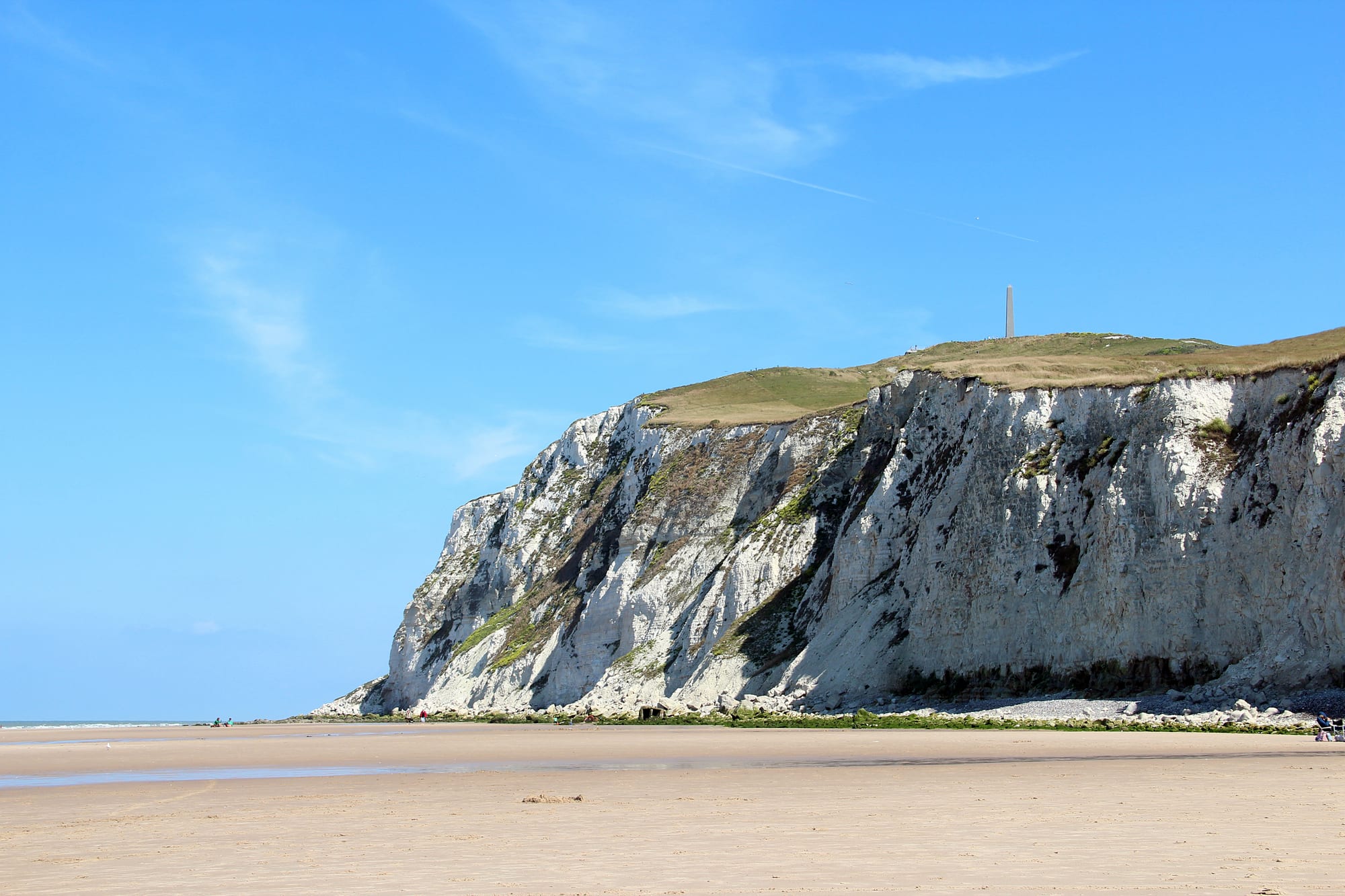 A white chalk headland topped with grass and a stone obelisk rising above a wide sandy beach under a blue sky