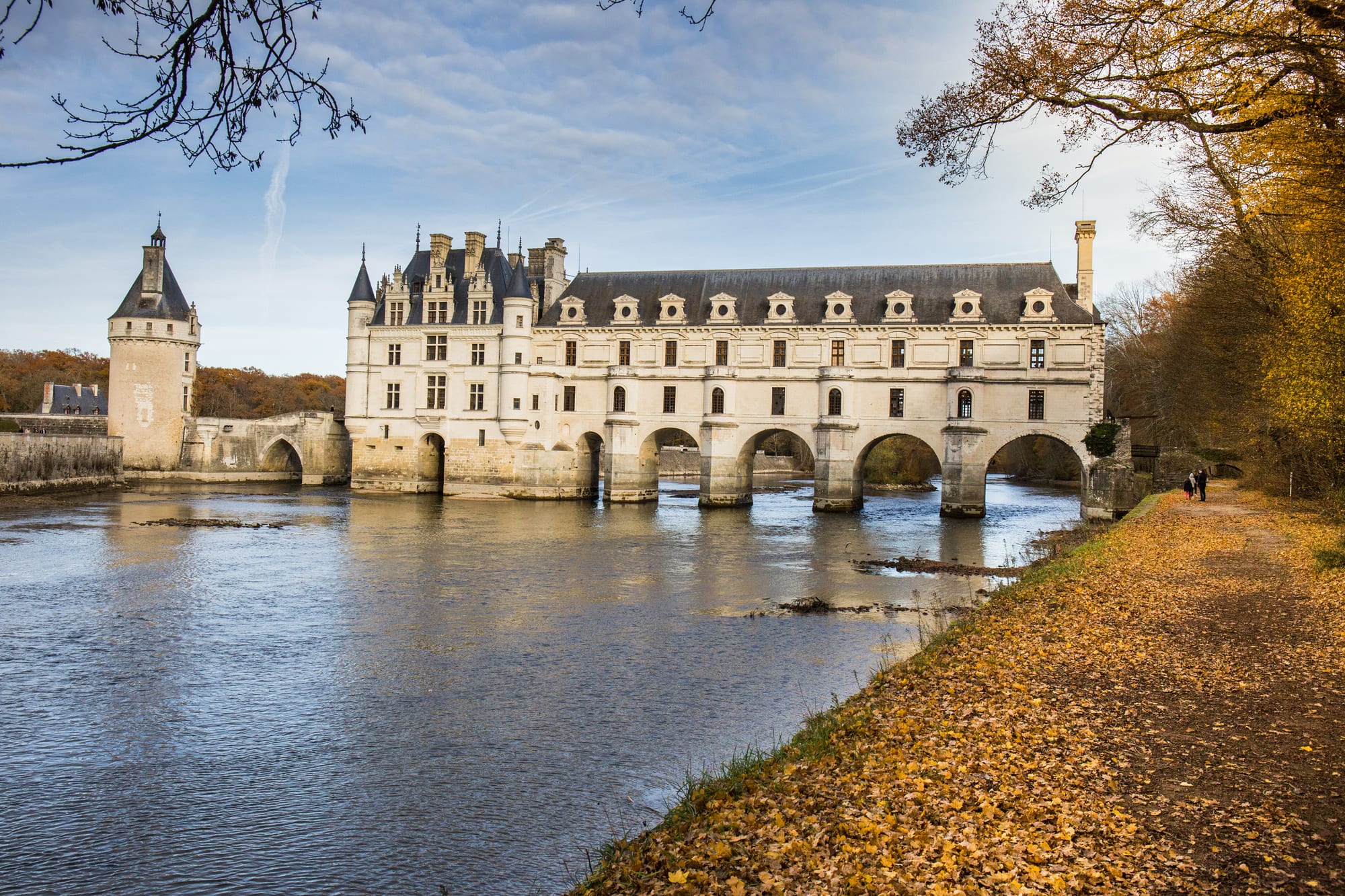 A white Renaissance château built on arches across a river, seen from a leaf-strewn riverside path in autumn
