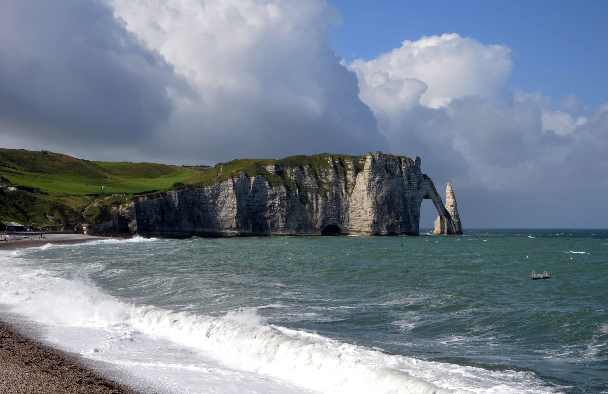 White chalk cliffs with a natural arch and a pointed sea stack above a choppy green sea under towering clouds
