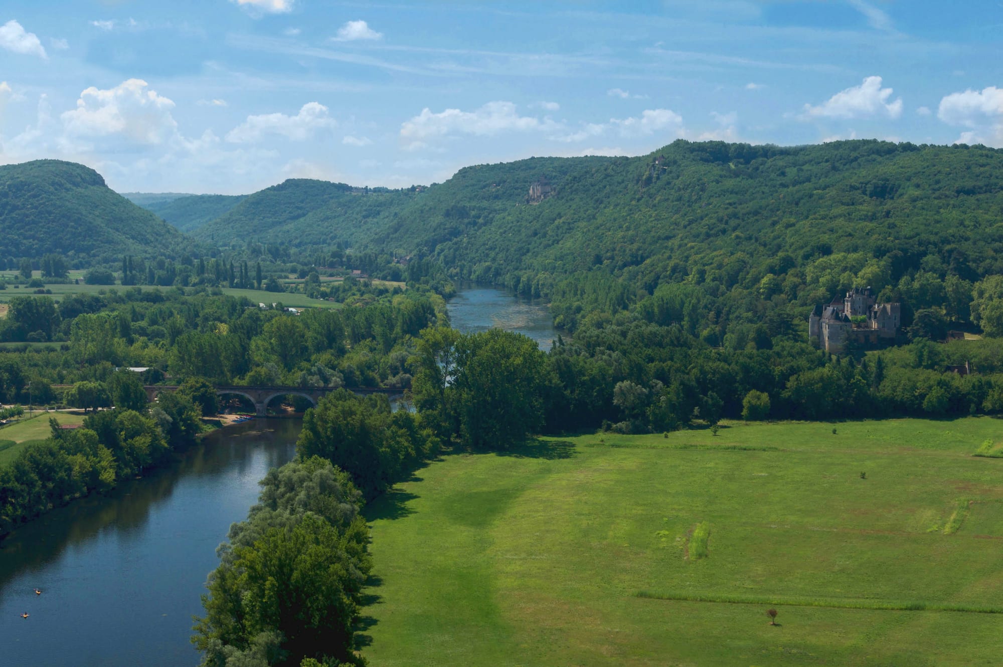 The Dordogne river winding between wooded hills and green meadows, with a stone bridge and a castle among the trees