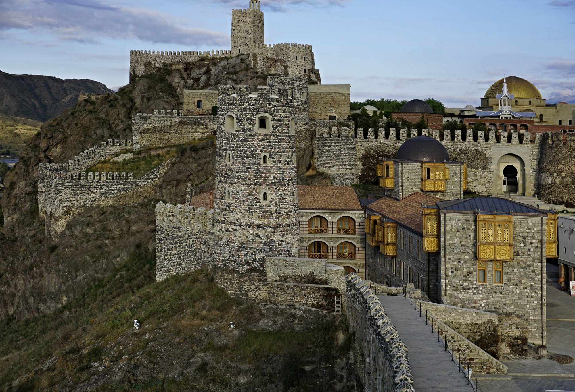 Stone fortress walls and towers climbing a rocky hill, with a gold-domed mosque and houses with ornate wooden balconies inside the walls