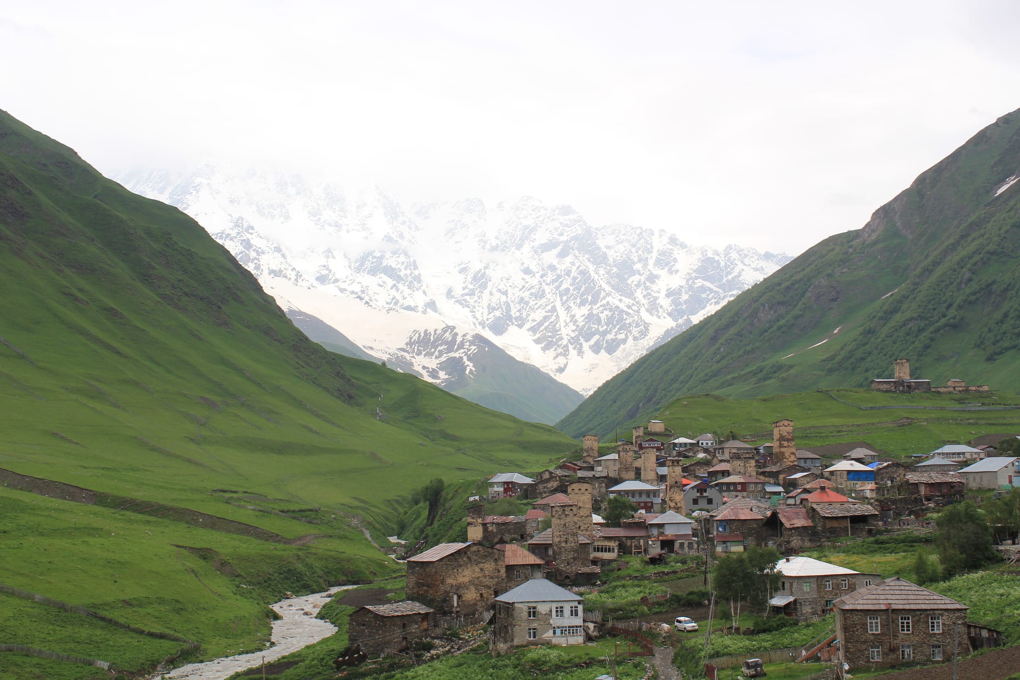 Stone tower-houses of a mountain village in a green valley, with a snow-covered massif rising into cloud behind