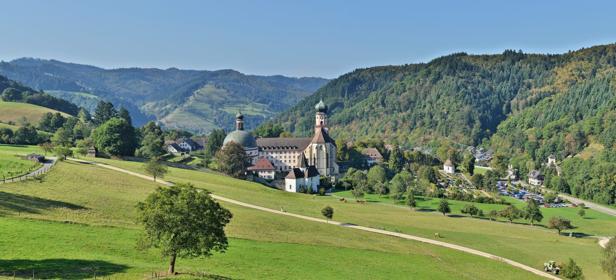 Green meadows and a winding path leading to a white abbey church with an onion-domed tower, set among wooded hills