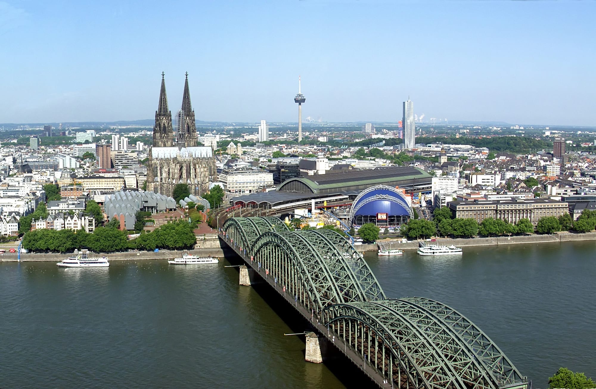 The green steel arches of a railway bridge crossing a wide river towards a twin-spired Gothic cathedral and a city skyline