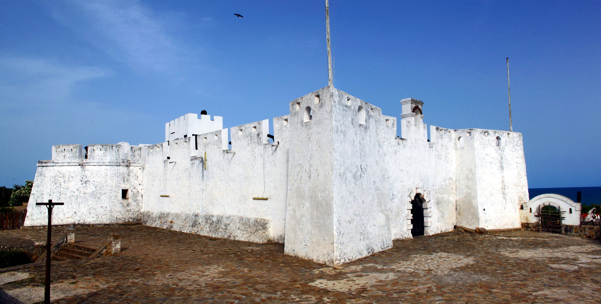 A whitewashed fort with crenellated walls and square bastions on a cobbled terrace under a deep blue sky, the sea visible at the edge