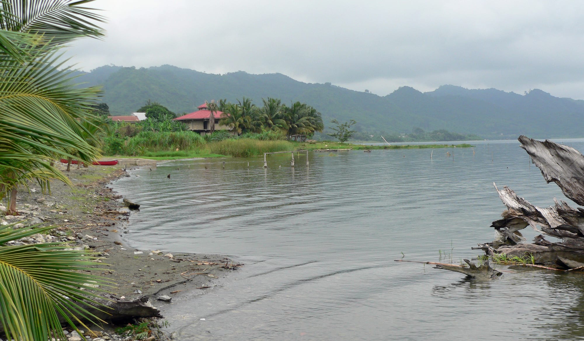 The calm grey water of Lake Bosomtwe with palm trees, a red-roofed house and forested crater hills under cloud