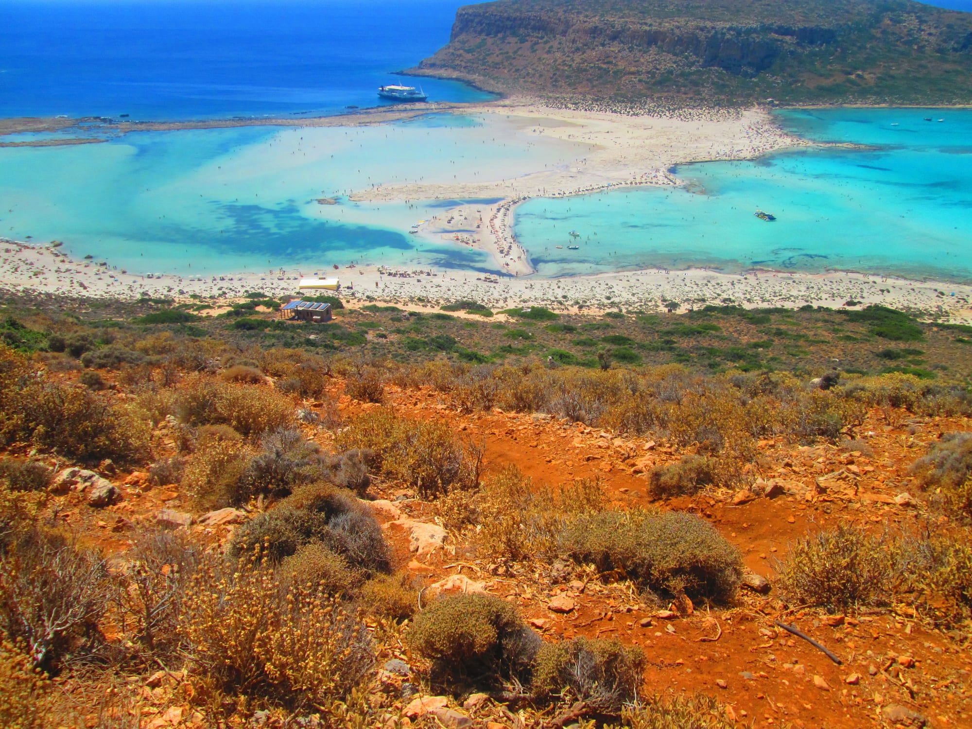 A turquoise lagoon and white sand spit seen from a hillside of red earth and low dry shrubs, with a rocky headland beyond