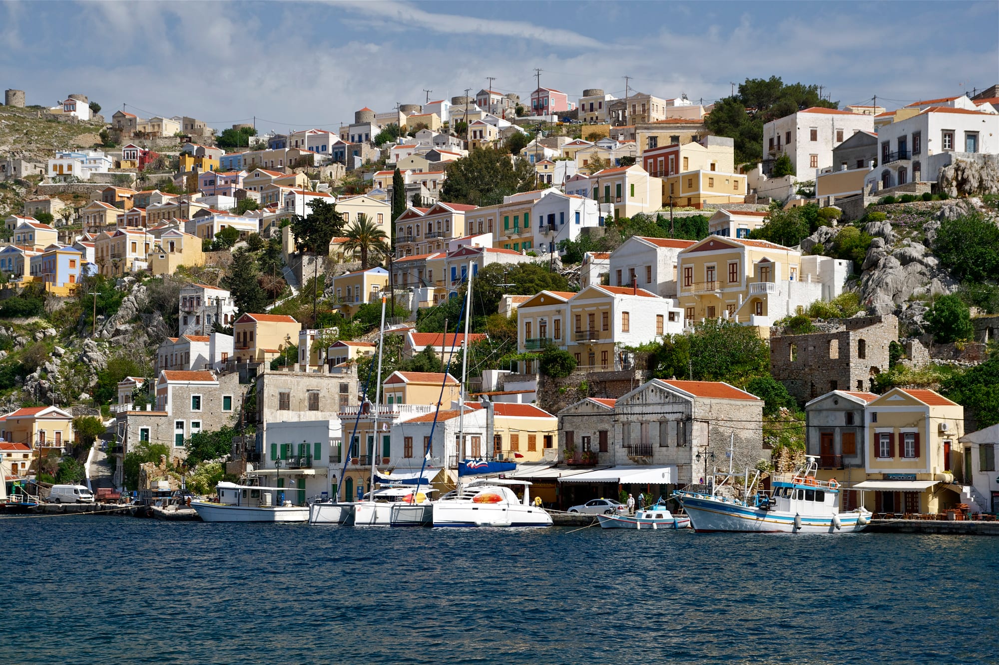 Pastel-coloured neoclassical houses with tiled roofs climbing a rocky hillside above a harbour lined with yachts and fishing boats
