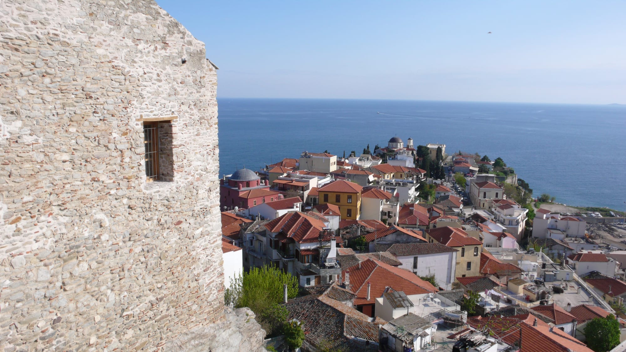 Red-tiled roofs and domes of an old town on a headland above a calm blue sea, seen past a rough stone castle wall