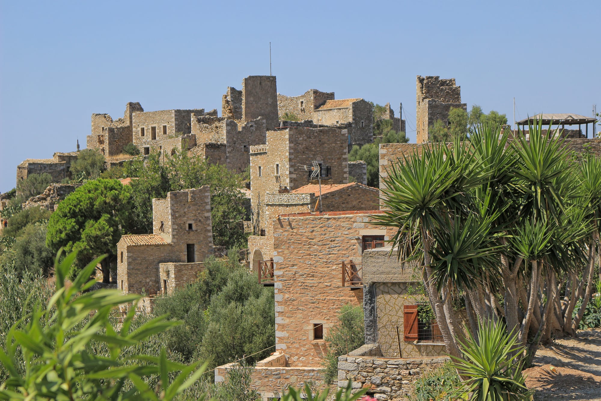 Square stone tower-houses clustered on a hilltop under a clear blue sky, with green trees and spiky yuccas in front