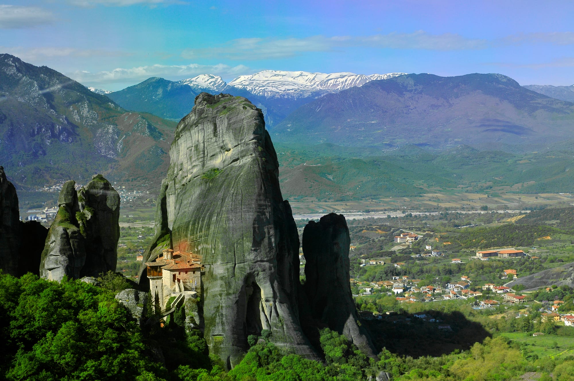 A red-roofed monastery on a ledge of a towering grey rock pinnacle above a green valley, with snowy mountains beyond