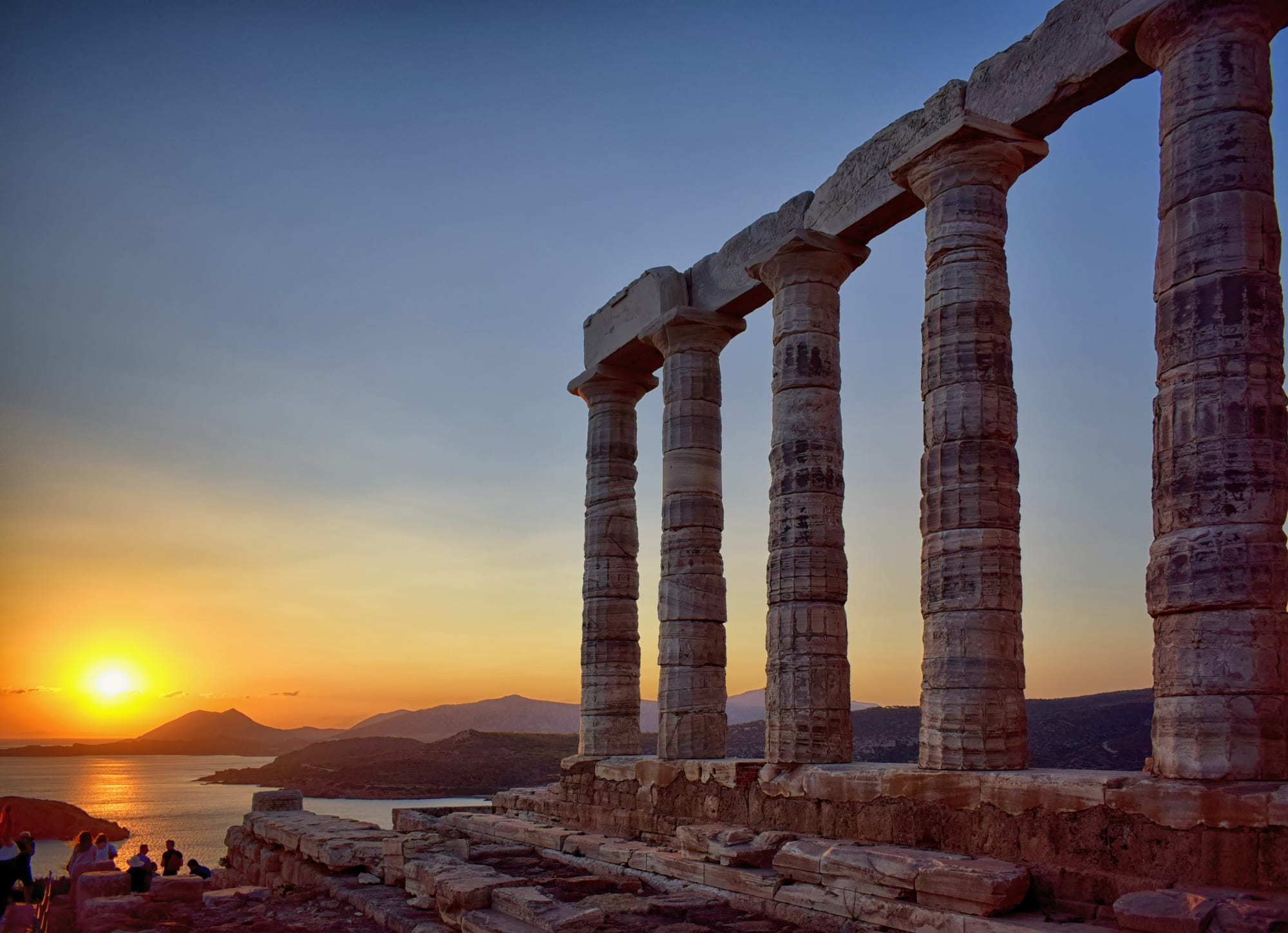 A row of weathered marble temple columns glowing in warm light as an orange sun sets over the sea and hazy blue headlands, with a few small figures watching from the rocks below