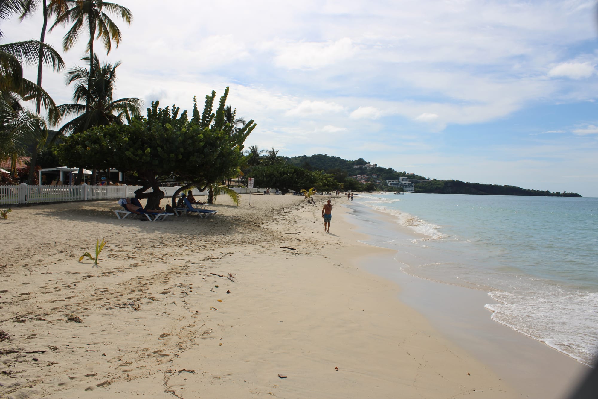 A long curve of pale sand meeting calm turquoise water, green hills and a few low buildings behind