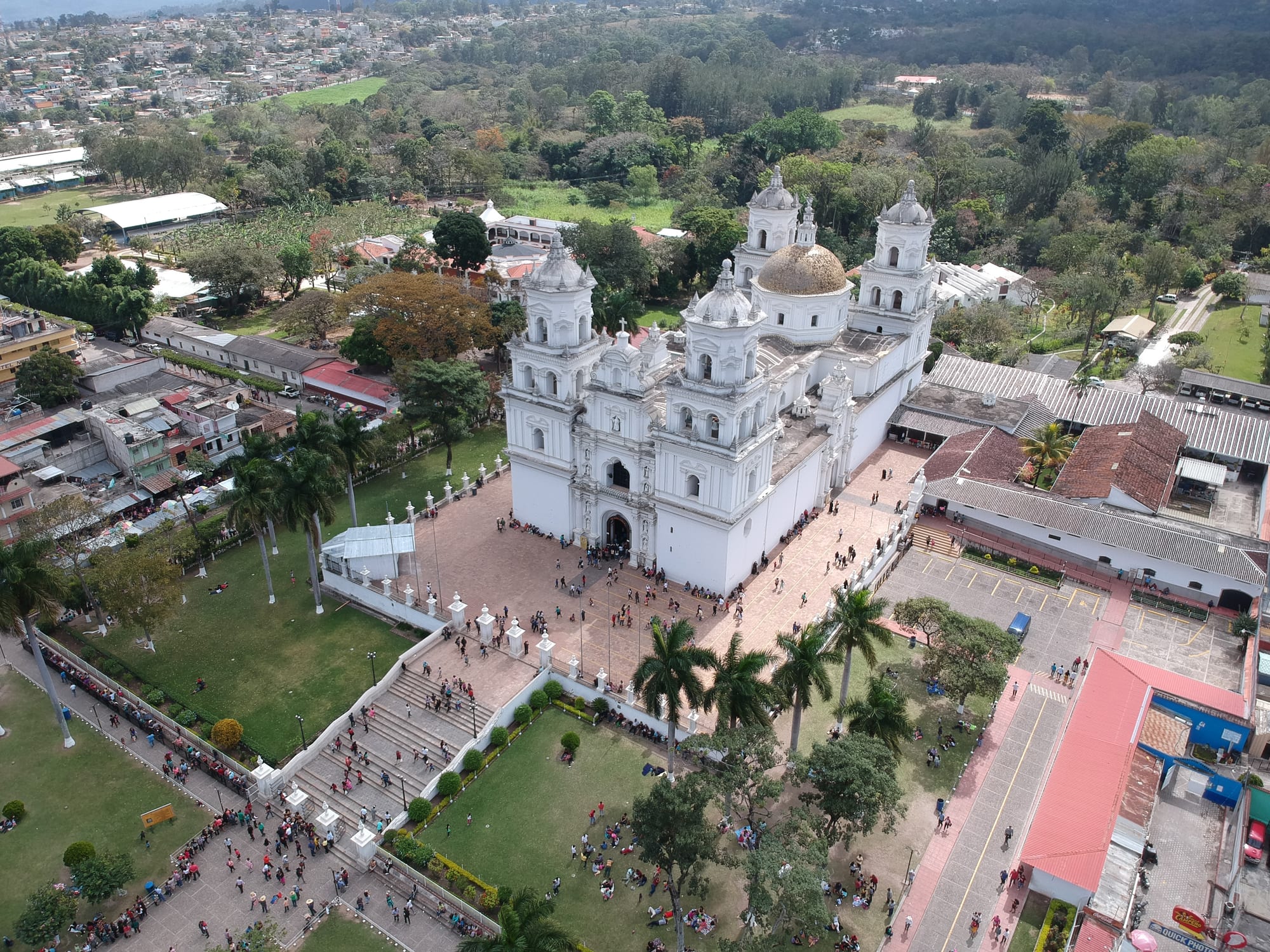 Aerial view of a large white baroque church with four towers and a dome, pilgrims crowding its steps and gardens