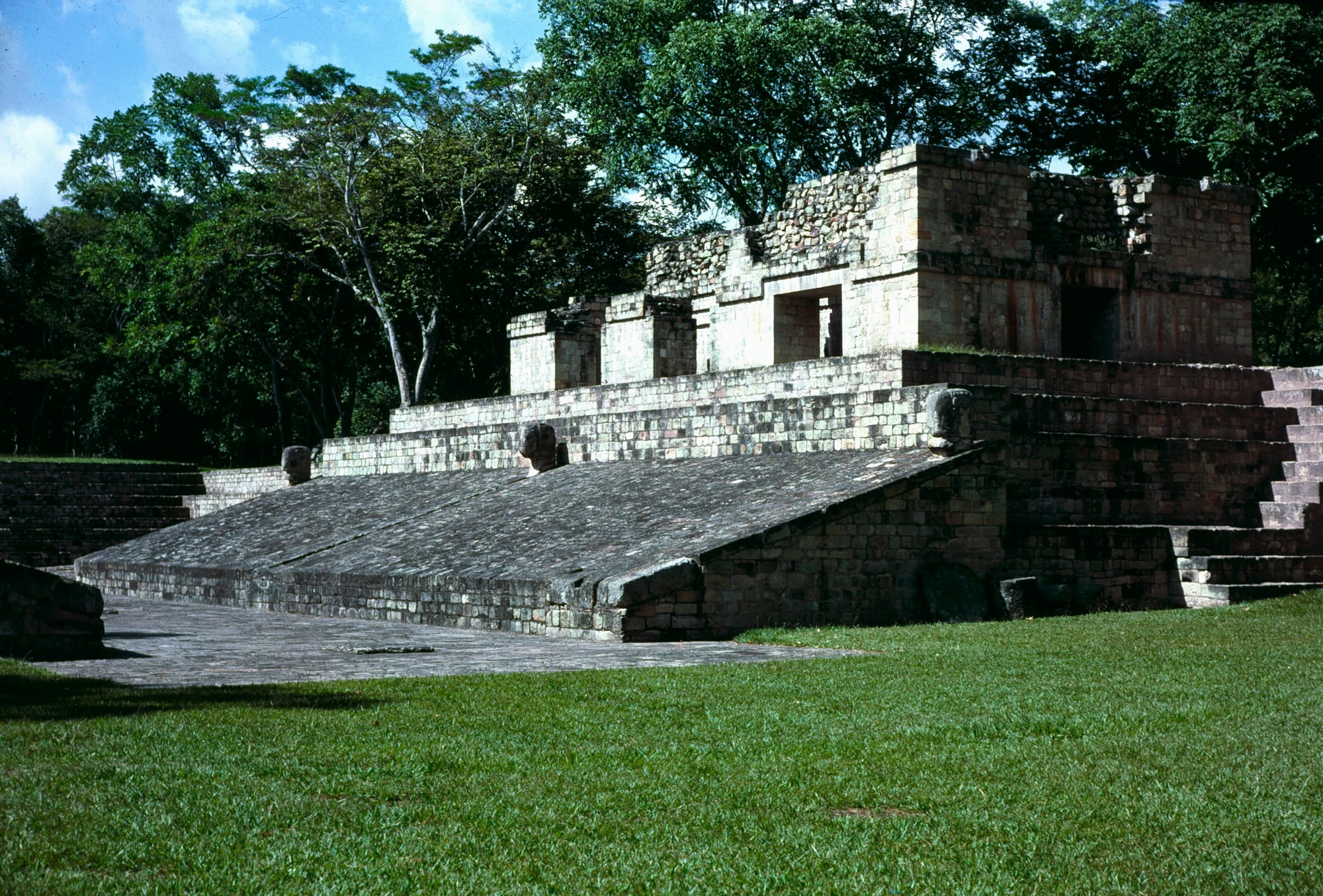 The sloping stone sides and upper buildings of a Maya ball court beside a lawn, backed by tall trees