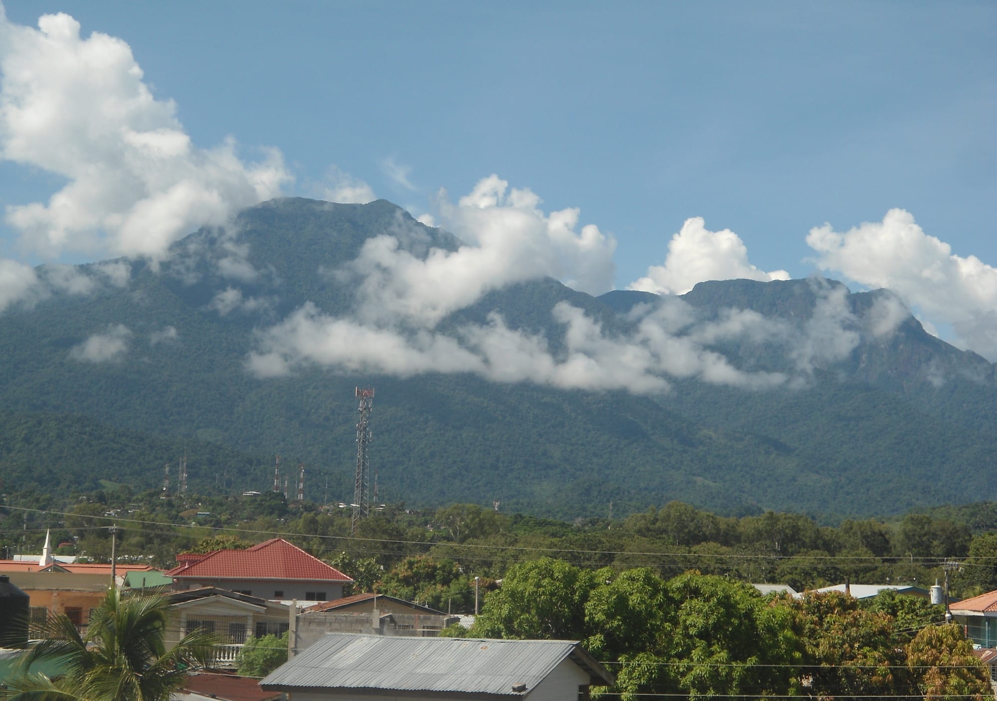 Forested mountains wreathed in low white cloud rising behind the red and tin roofs of a town