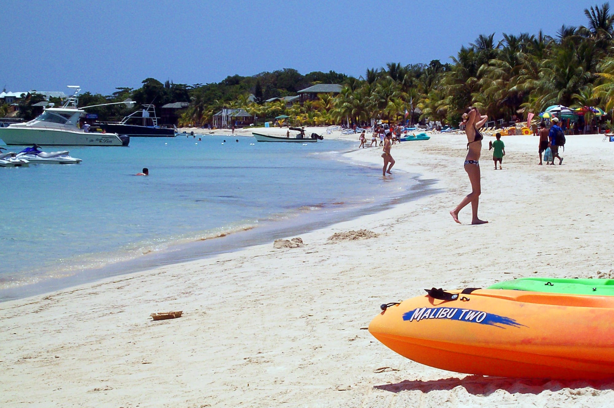 A curve of white-sand beach backed by palms, with turquoise shallows and a reef line offshore under a bright sky