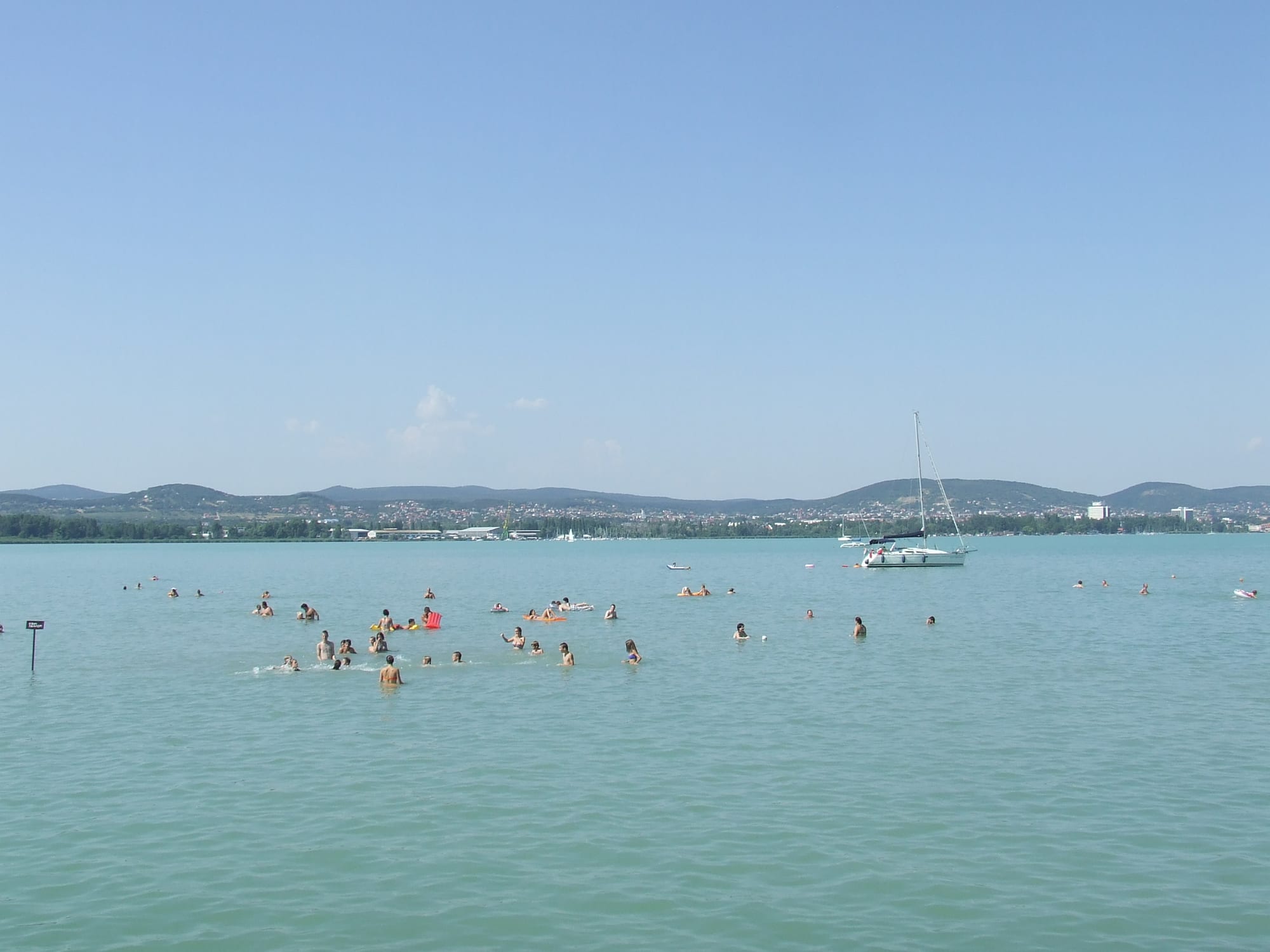 Swimmers and a moored sailing boat in pale green lake water, with low wooded hills and a town on the far shore