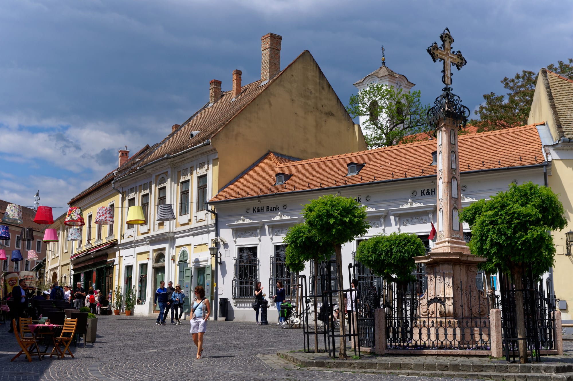 A cobbled square lined with pastel baroque houses with tiled roofs, a tall ornate stone cross in a railed enclosure and a church tower behind