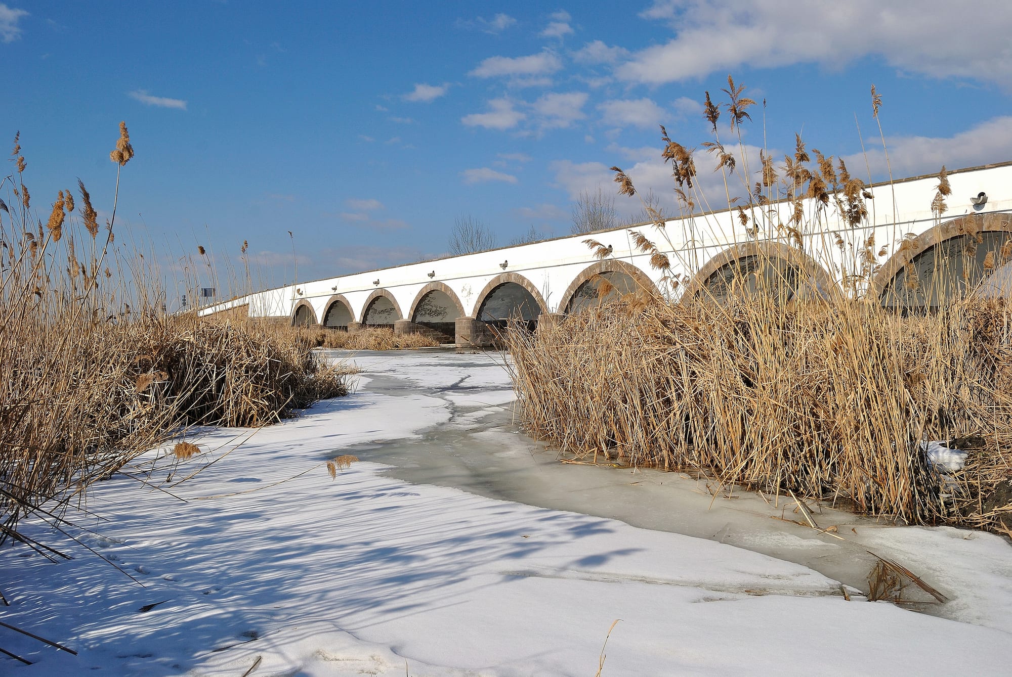 A long white stone bridge with arched openings over a frozen, snow-covered stream lined with dry reeds