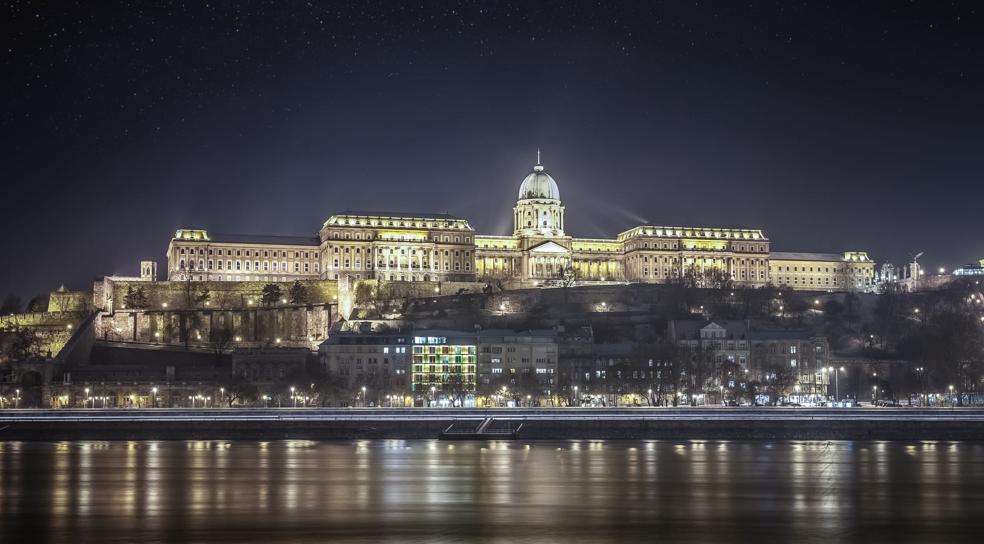 A huge floodlit palace with a green central dome on a fortified hill at night, its lights reflected in a wide river under a starry sky