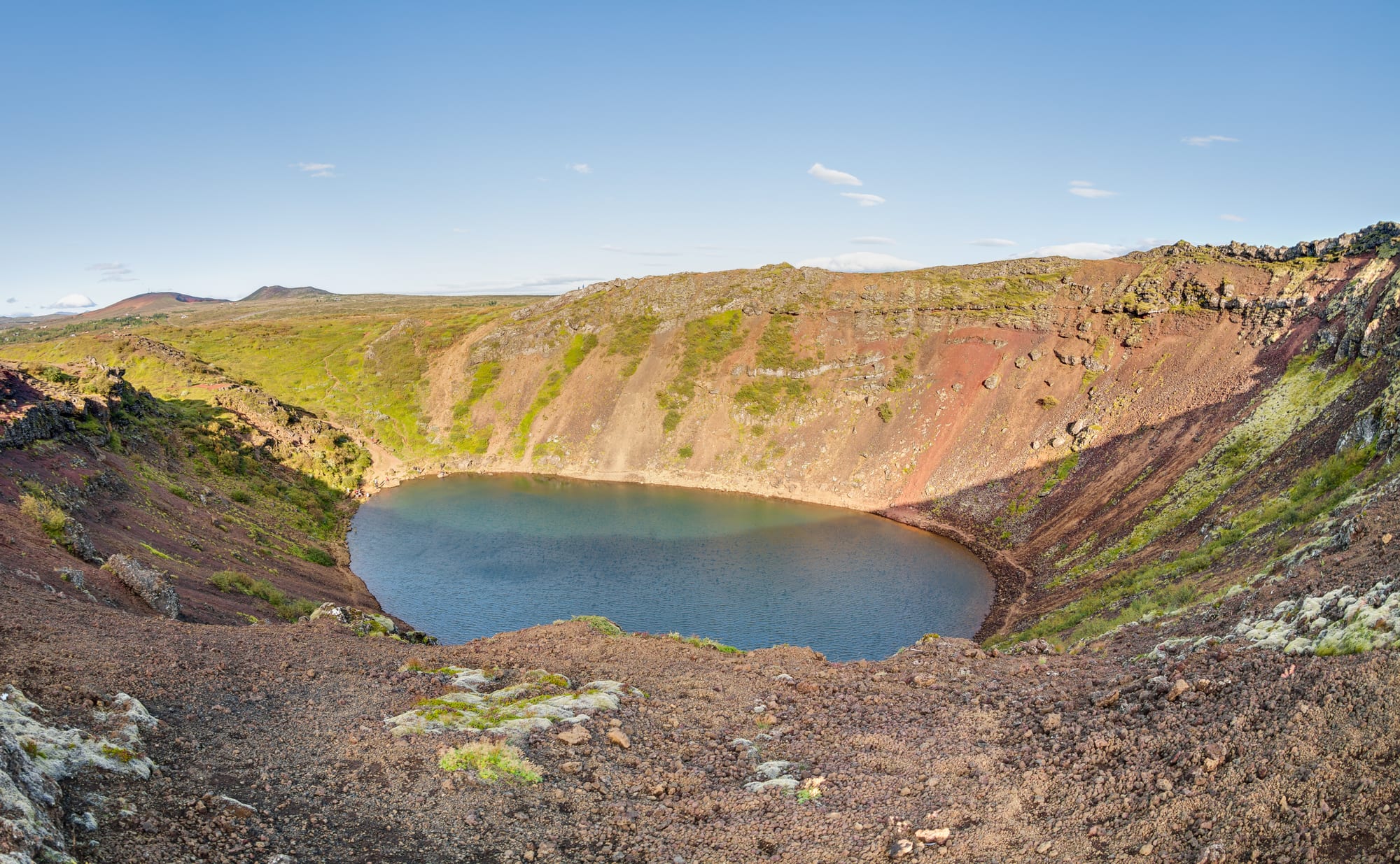 A still blue-green lake at the bottom of a volcanic crater with steep red and brown slopes patched with moss, under a clear sky