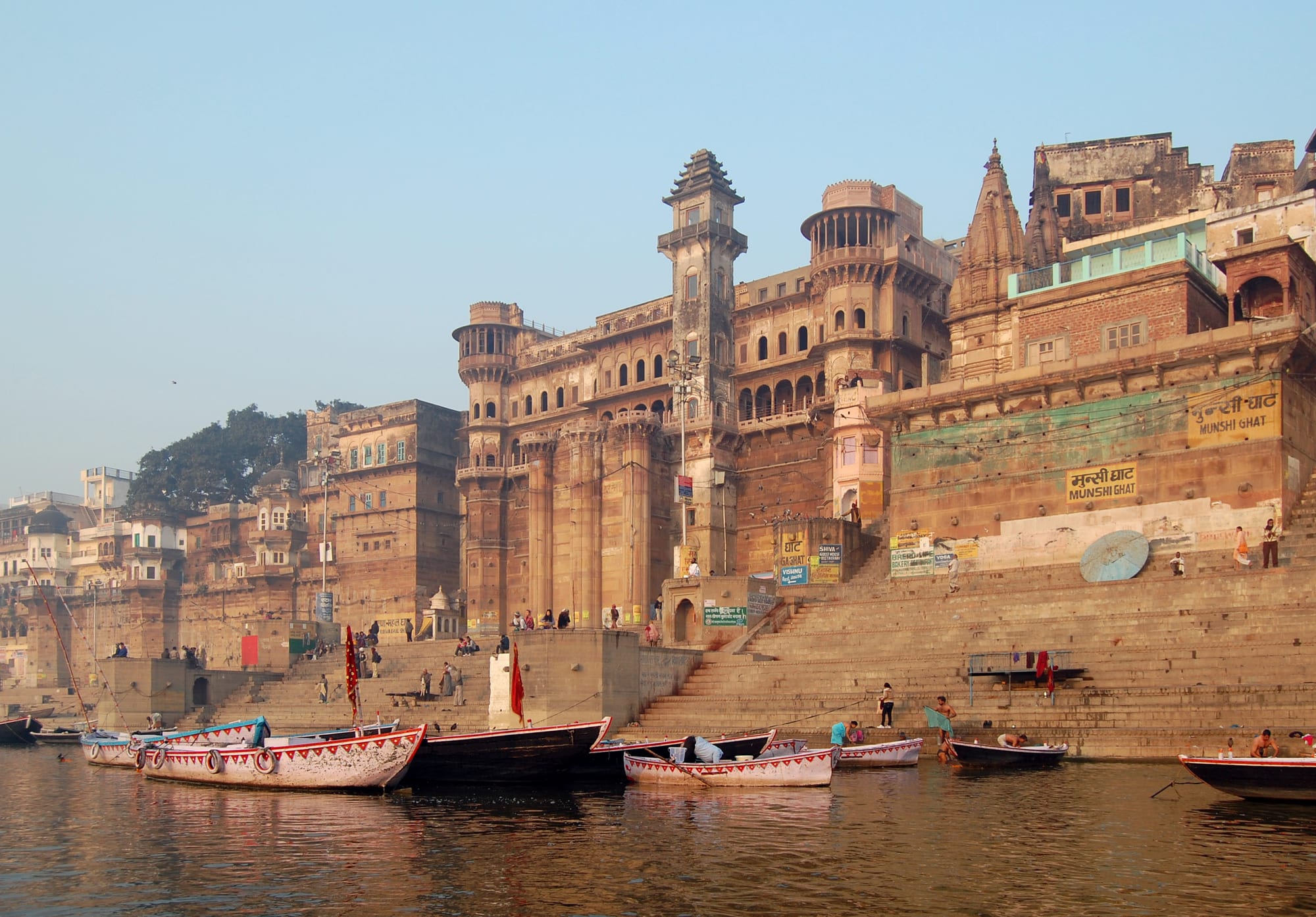 Sandstone palaces and towers rising above broad stone steps on a riverbank, with wooden boats moored in the water