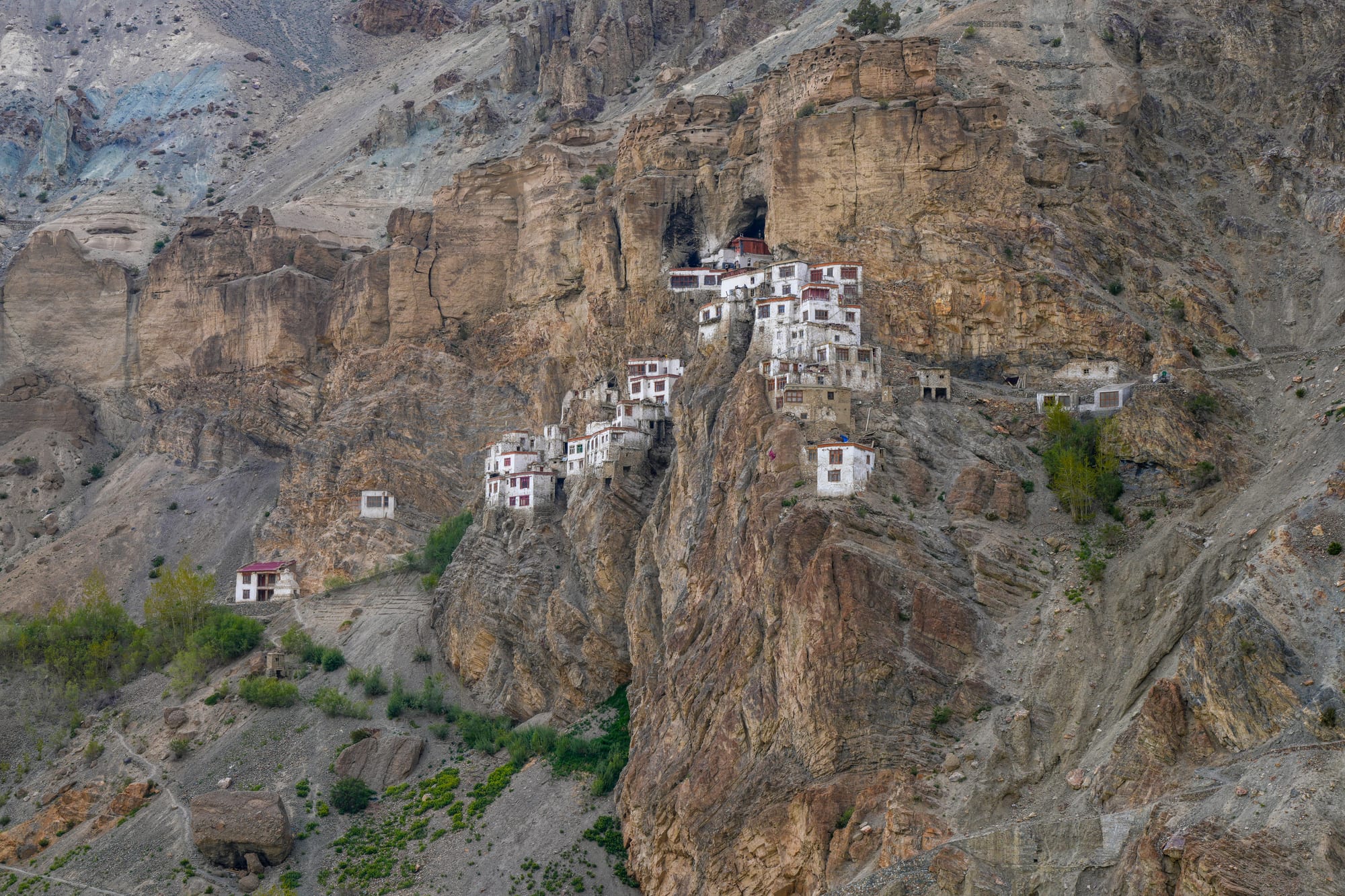 White monastery buildings clinging to a cave-topped cliff face above a rocky gorge, with patches of green below