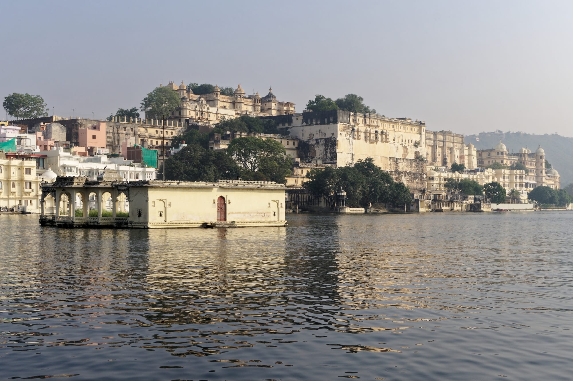 Cream-coloured palace buildings and domes above the calm water of a lake, with a small pavilion in the foreground