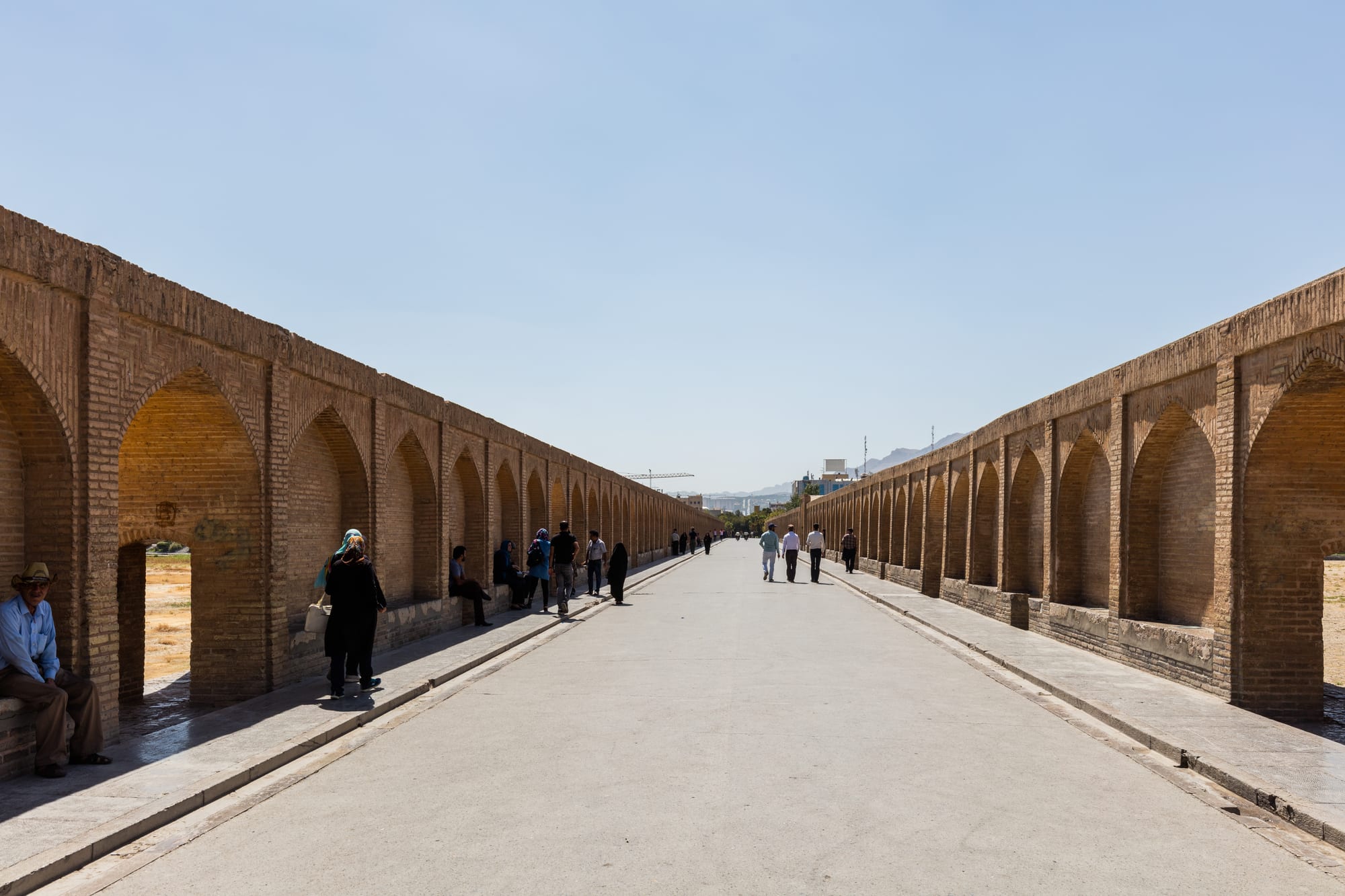 A long pedestrian bridge deck lined on both sides by rows of brick arches, with people walking and sitting in the shade