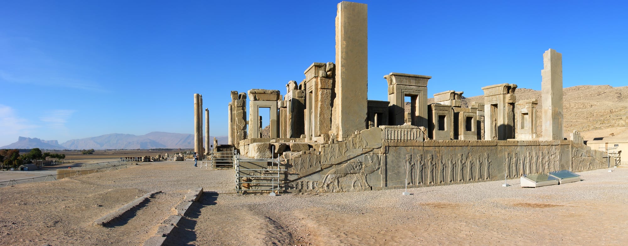Stone doorways and columns of an ancient palace above a staircase carved with rows of relief figures, under a clear sky