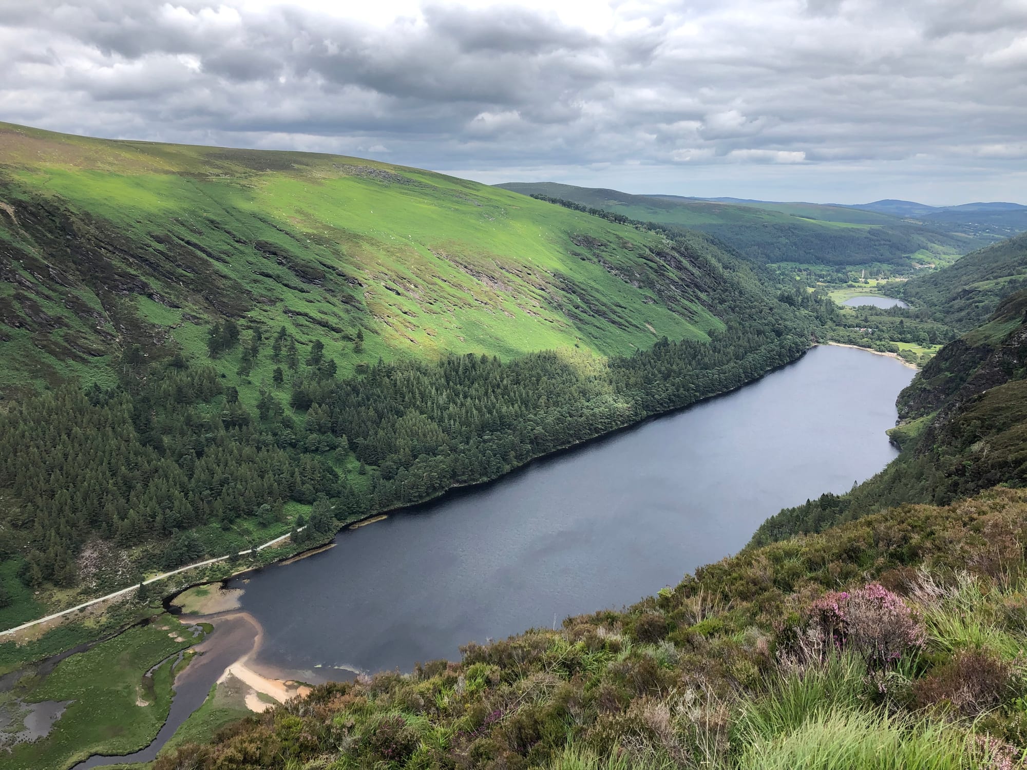 Long dark lake in a green glacial valley, forested slopes and heather in front, a smaller lake in the distance
